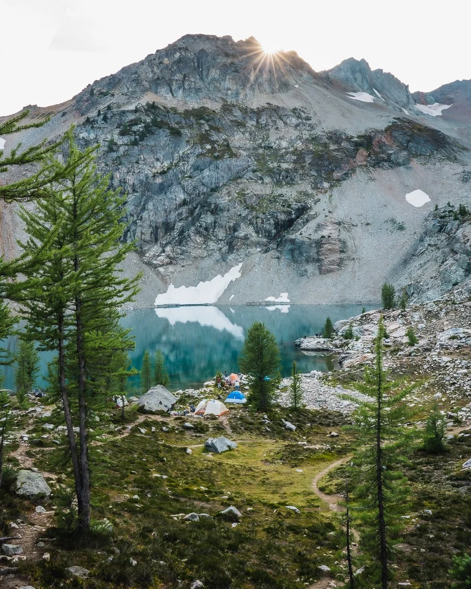 Scenic mountain landscape with a turquoise lake, evergreen trees, rocky terrain, and a group of campers near the water with tents, with snow patches on the mountains and a bright sun in the sky.
