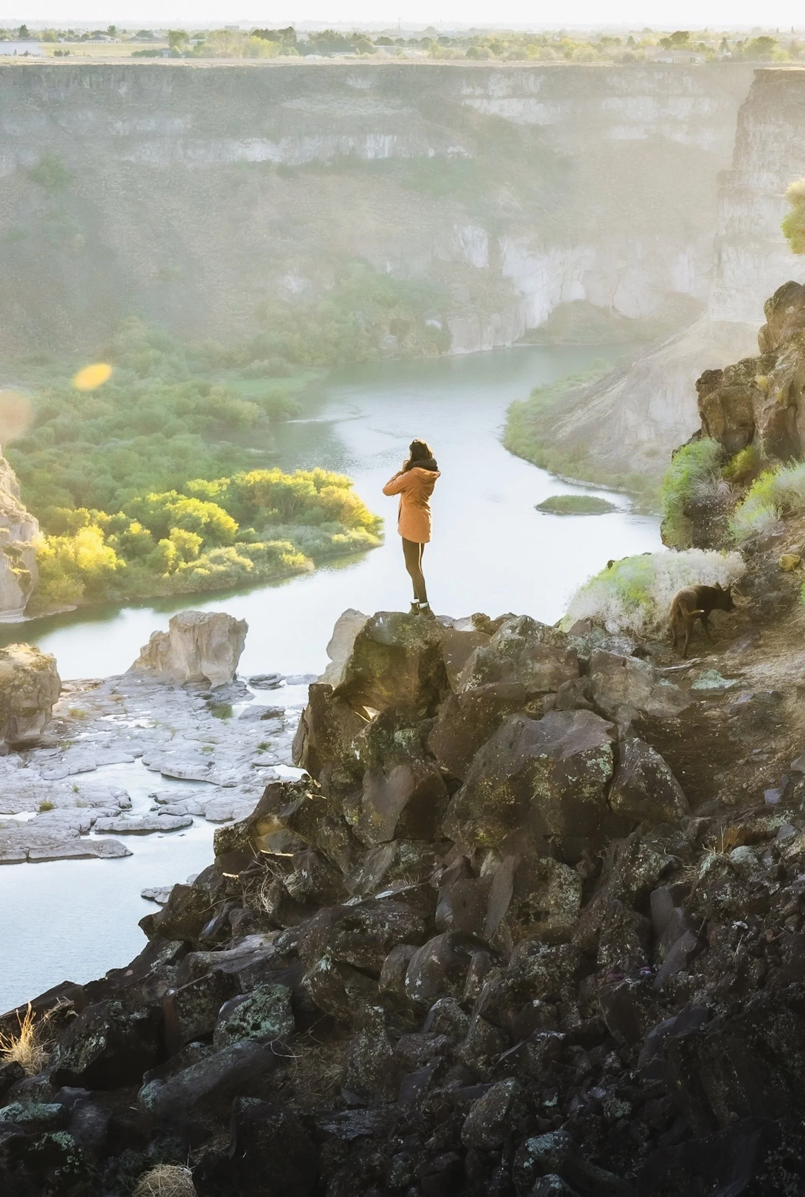 A woman standing on large rocks overlooking a river canyon with waterfalls and lush green trees, at sunrise or sunset.