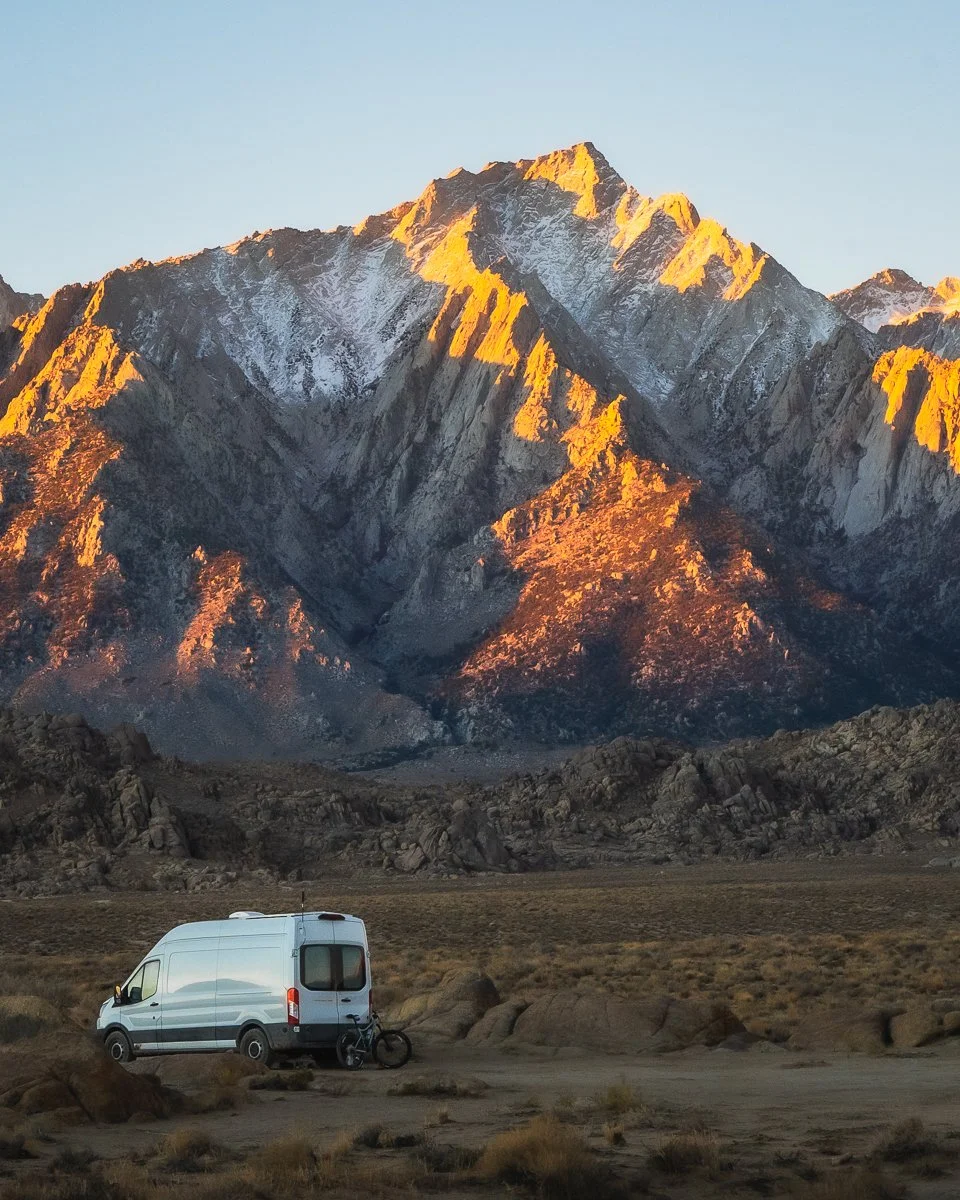 A white camper van and a bicycle parked on a dirt area in front of a mountain range at sunset, with snow-capped peaks illuminated orange by the setting sun.