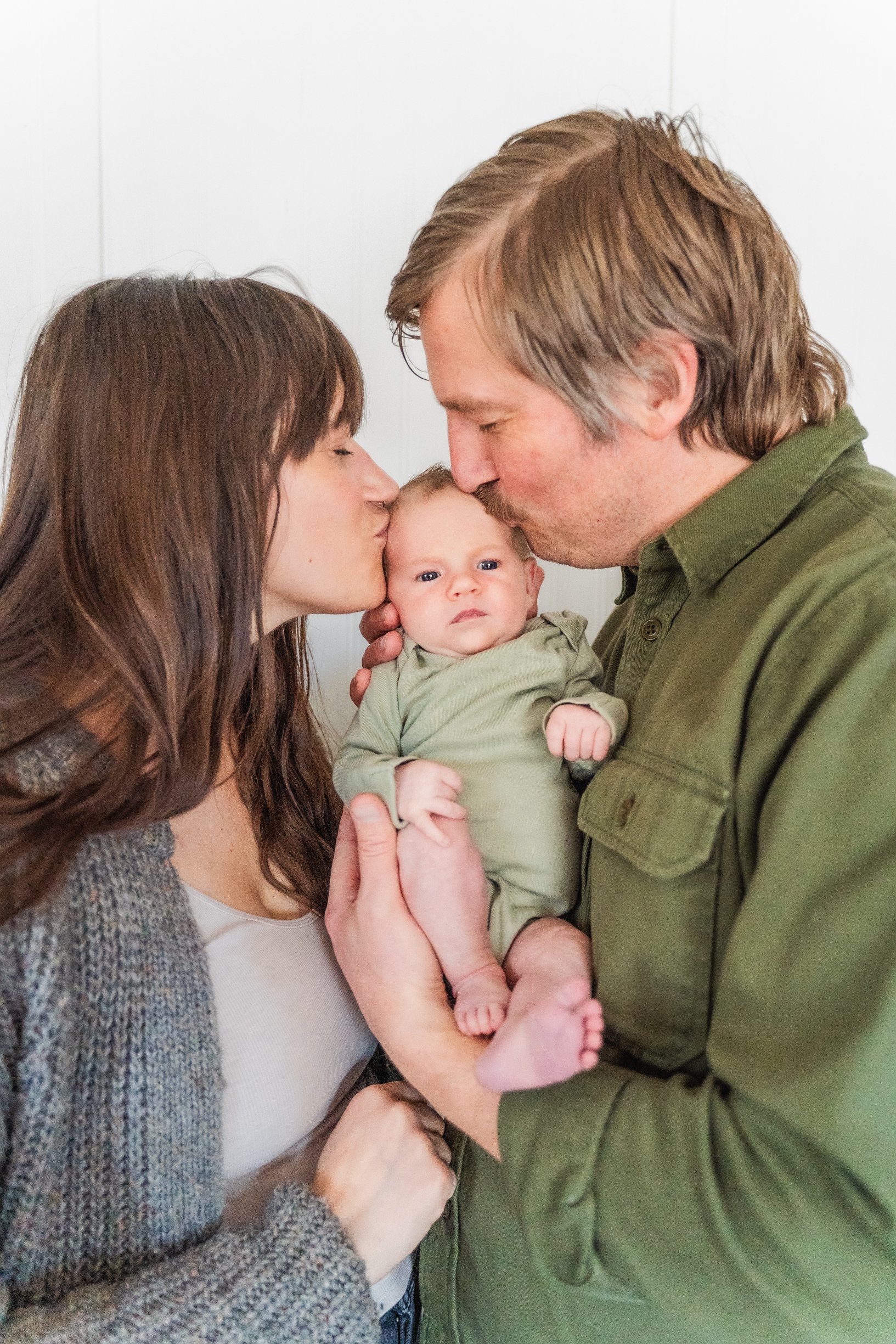 A family of three, a woman and a man, holding a baby between them. The woman and man are kissing the baby's head, and the baby is looking at the camera with a serious expression.