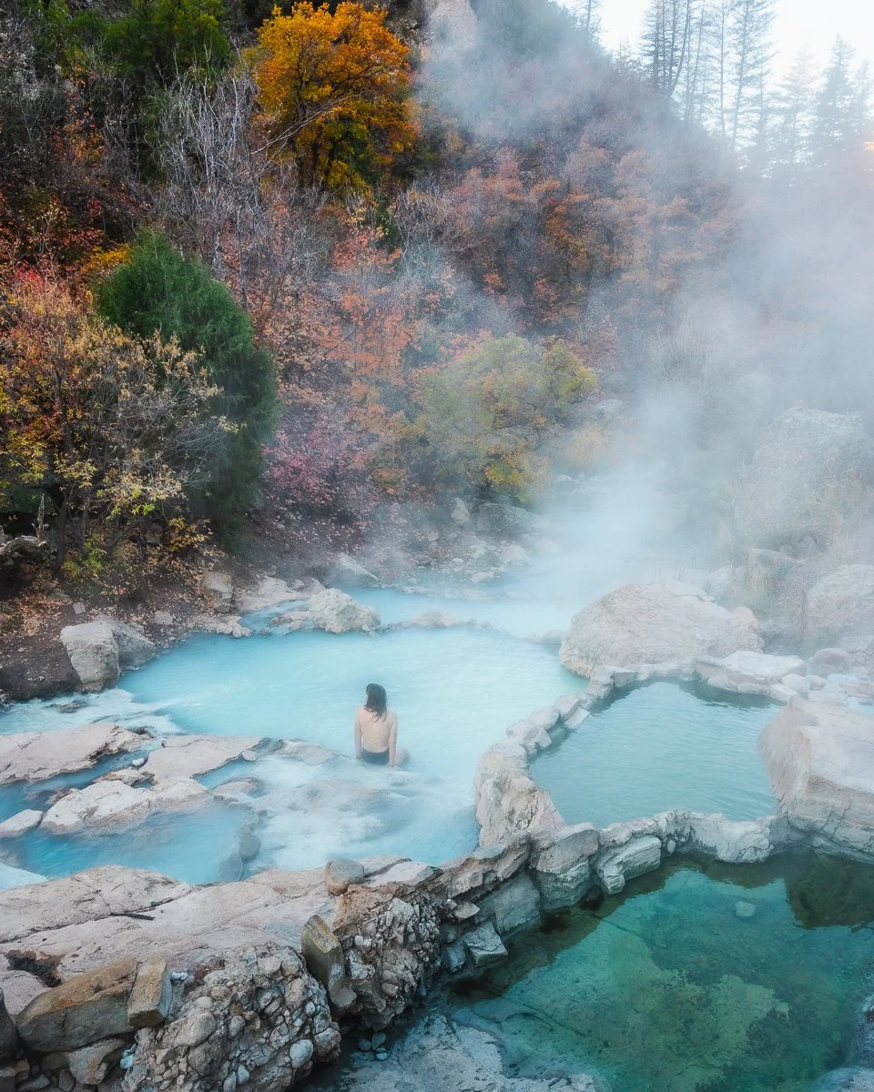 A woman sitting in a steaming hot spring with blue water, surrounded by rocks and trees with autumn foliage, on a misty day.