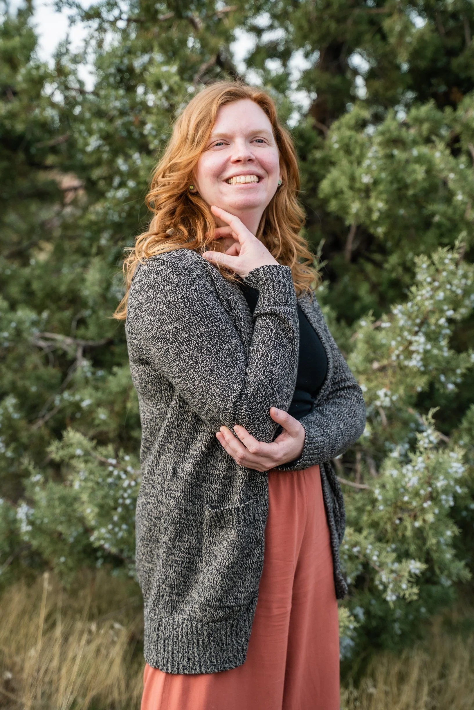 A woman with red hair smiling outdoors, standing in front of trees and foliage.