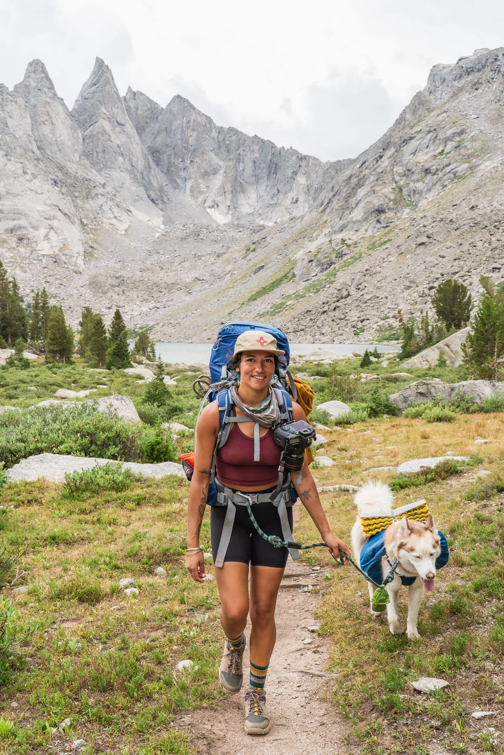 A woman hiking in a mountainous area with a husky dog, carrying a backpack and camera, on a trail surrounded by greenery and rocks.