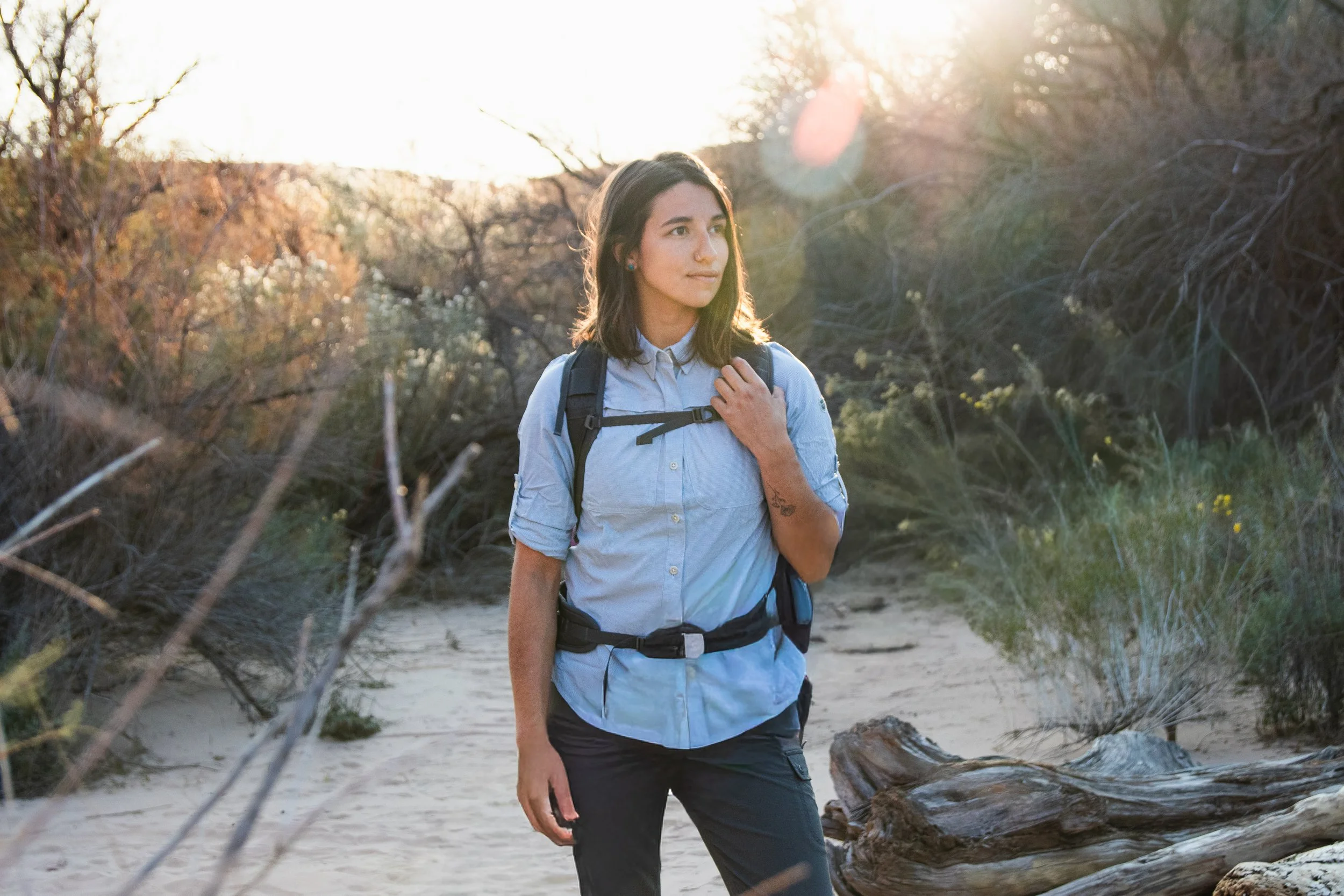 A woman with dark hair is walking outdoors on a trail in a natural setting during sunset. She is wearing a light blue button-up shirt, black pants, and a backpack, and she is looking to her left with a contemplative expression.