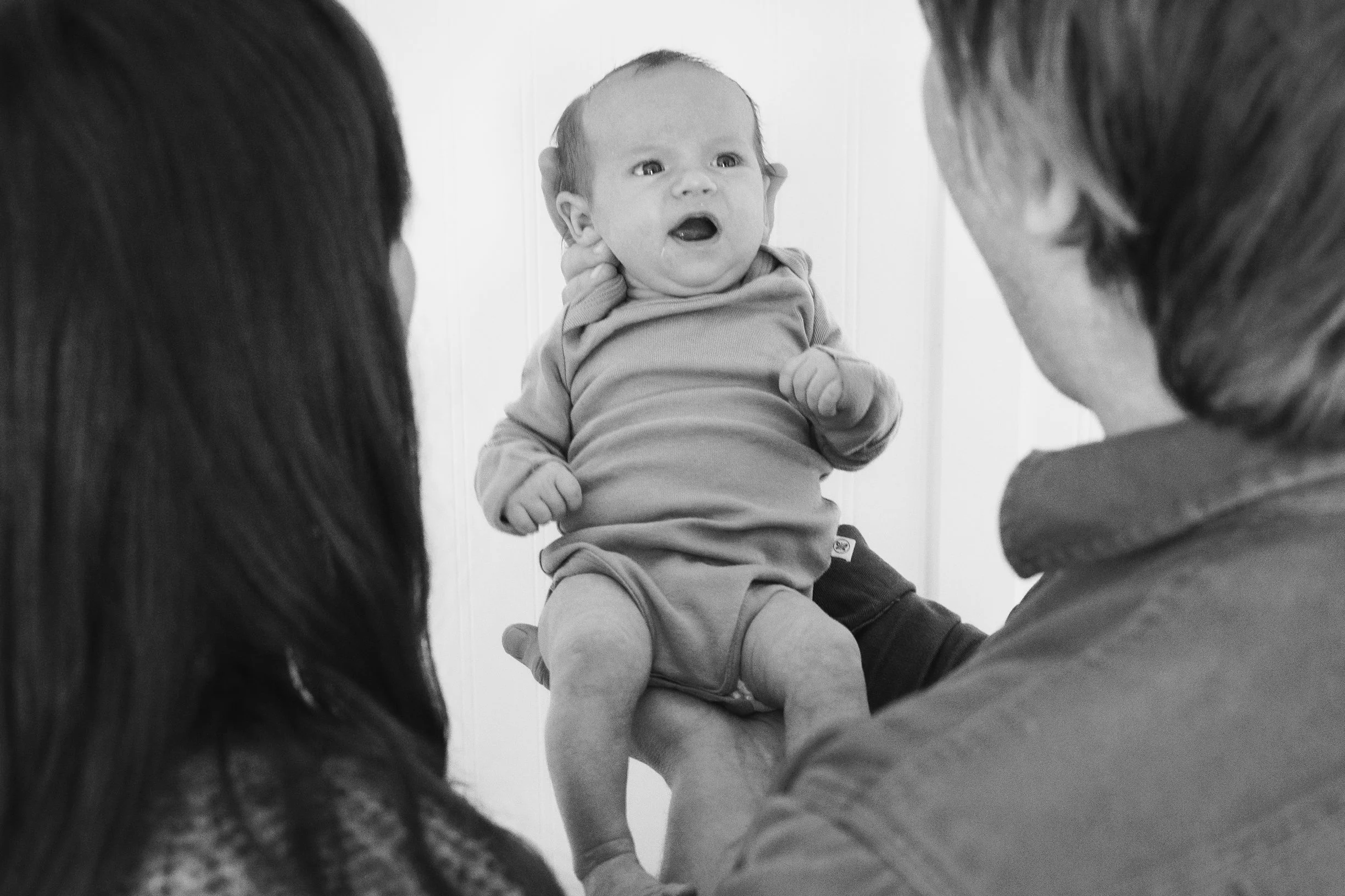 A baby being held up by a man while a woman looks on, with all faces visible in this black-and-white photo.