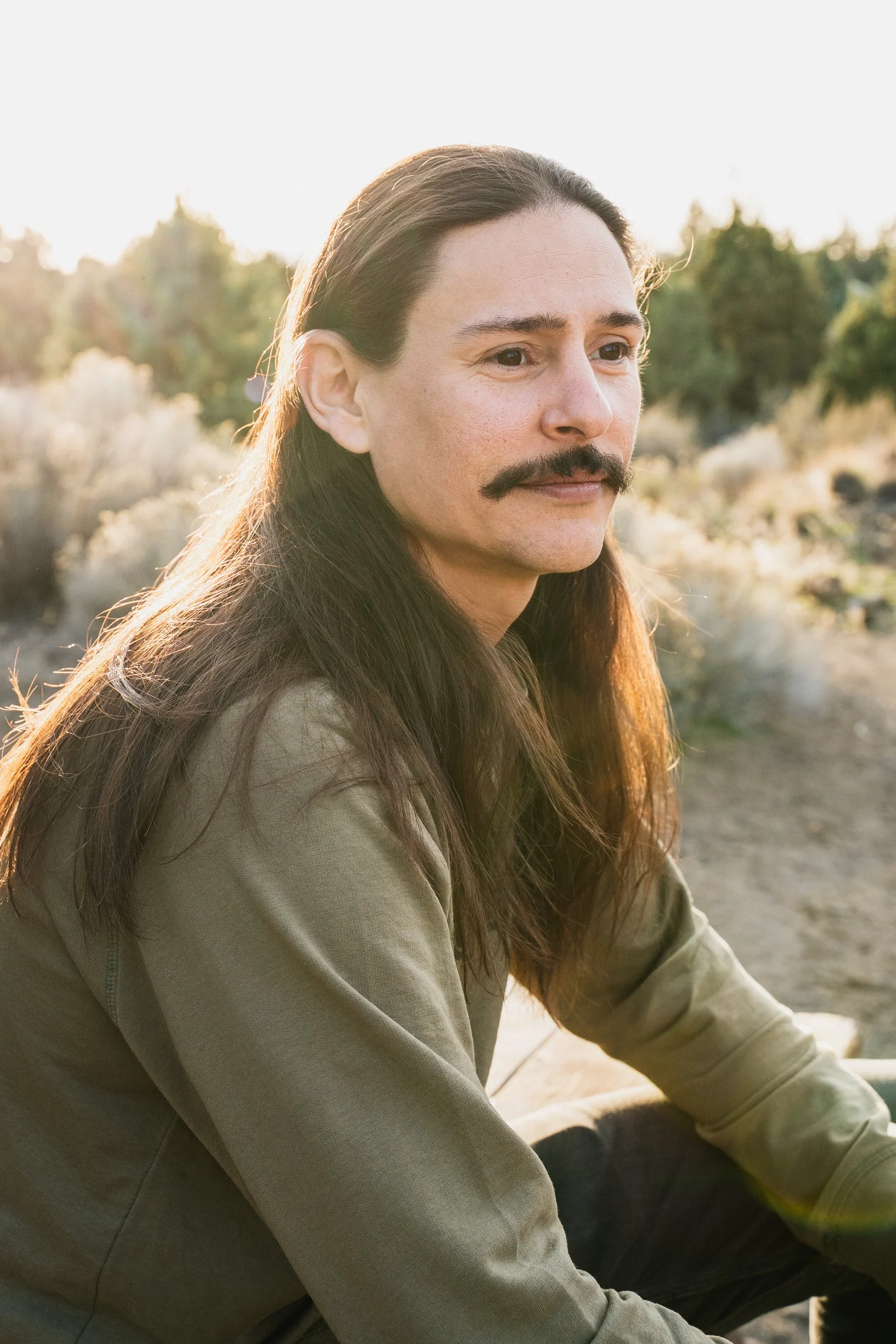 A man with long dark hair, a mustache, and a light olive jacket sitting outdoors during sunset, with a natural landscape background.