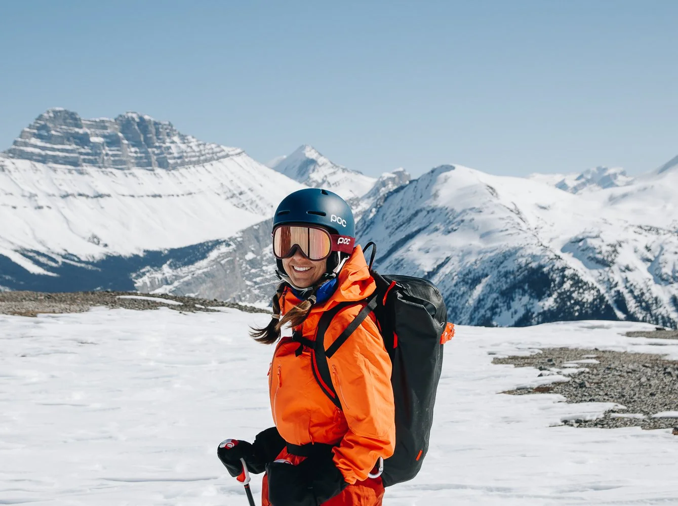 A woman in an orange jacket and black helmet, wearing snow goggles, stands on snowy terrain with snow-covered mountains in the background.