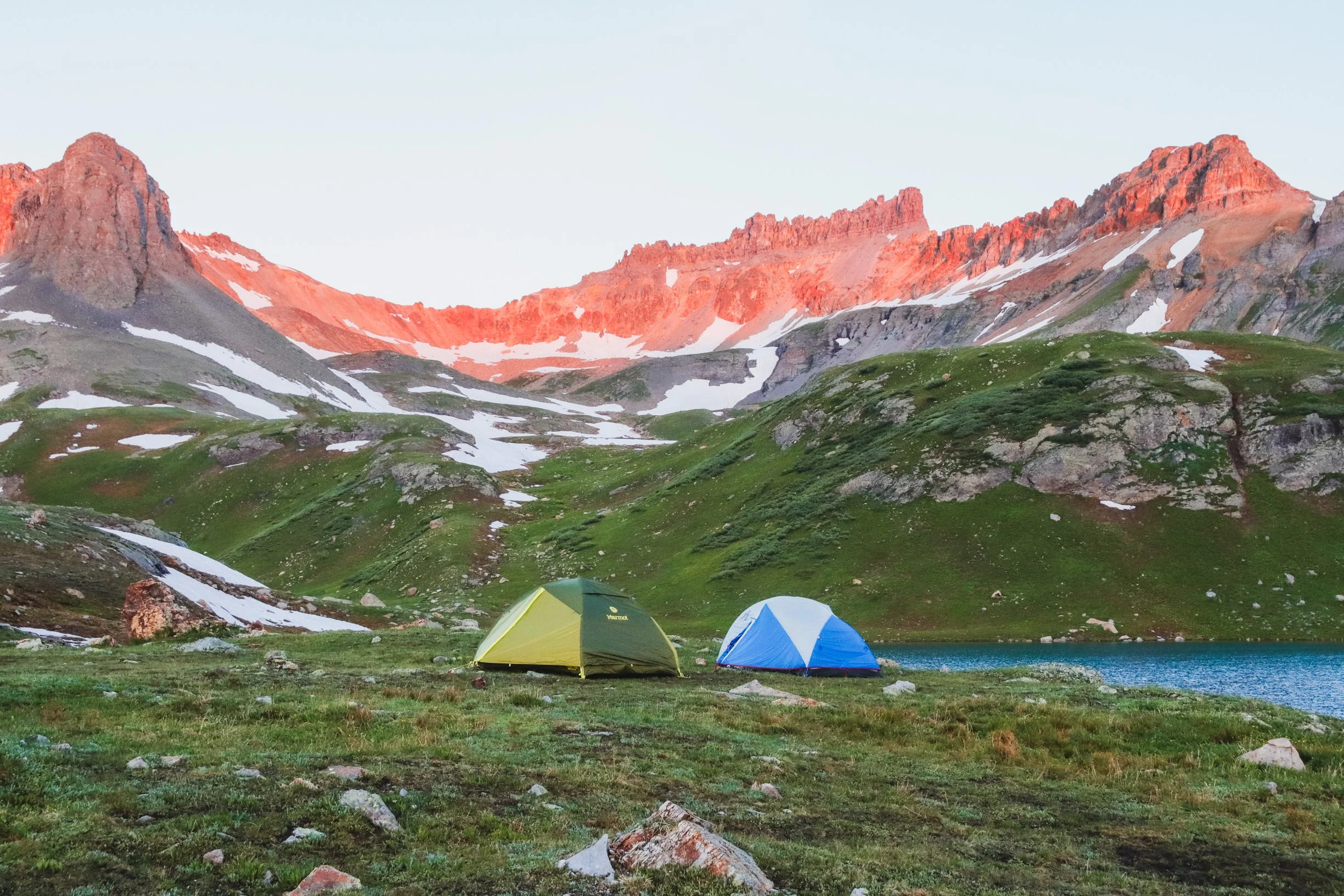 Two tents set up on a grassy area near a lake, with snow-capped mountains and pink-tinged peaks in the background during sunset or sunrise.