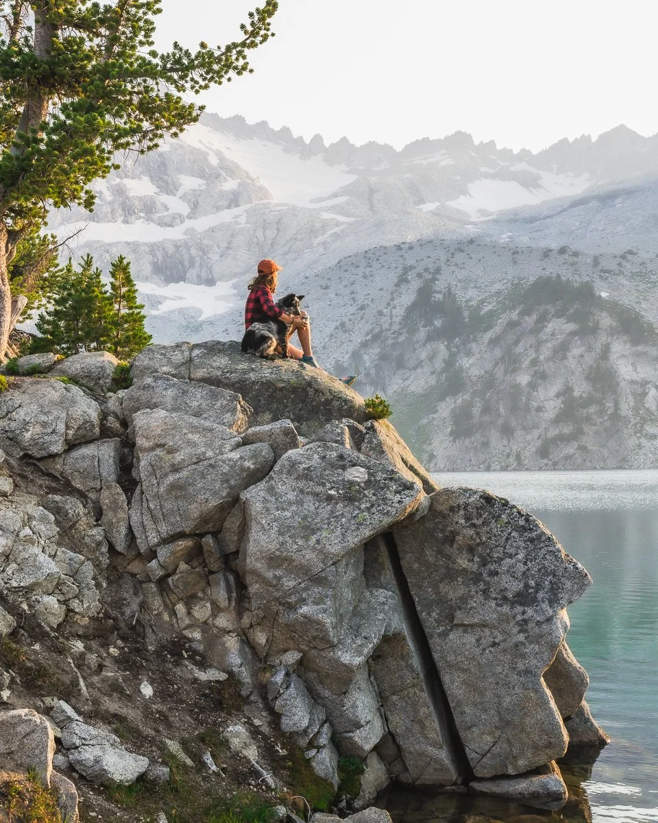 A person in a red plaid shirt and orange hat sitting on a rocky overlook with a dog, looking at a mountain lake with snowy peaks in the background.