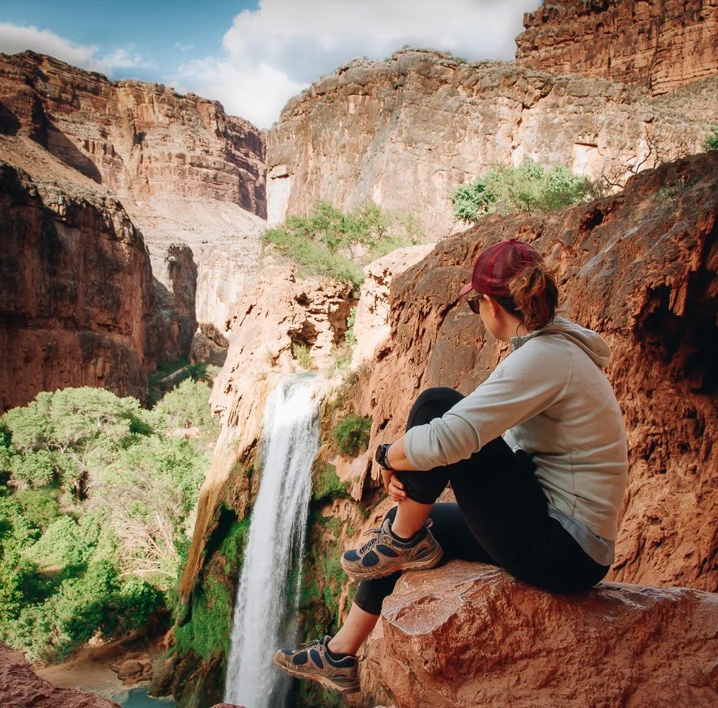 A woman sitting on a rocky ledge watching a waterfall in a canyon surrounded by tall red cliffs and green trees.
