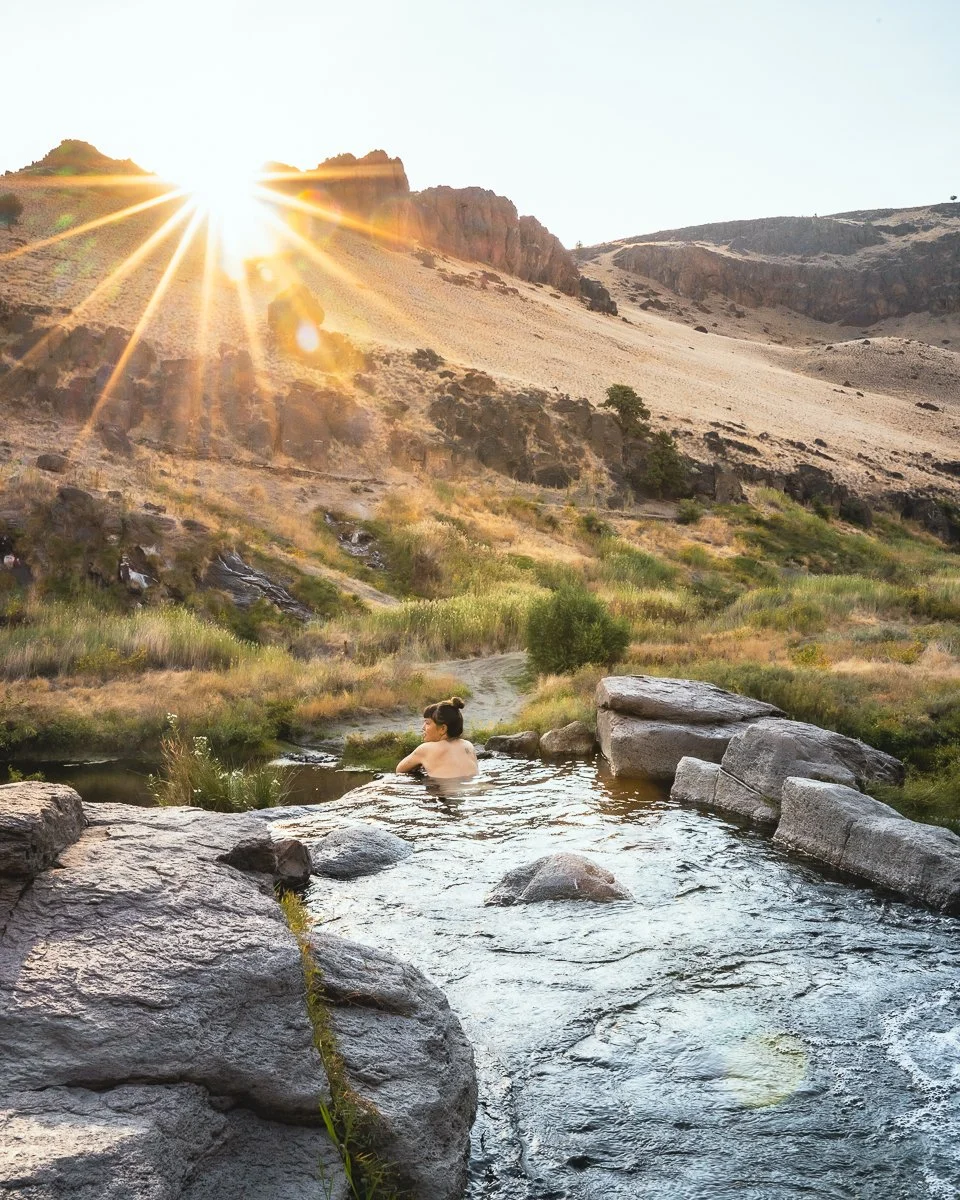 A woman relaxing in a hot spring surrounded by rocks, with a mountainous landscape and setting sun in the background.