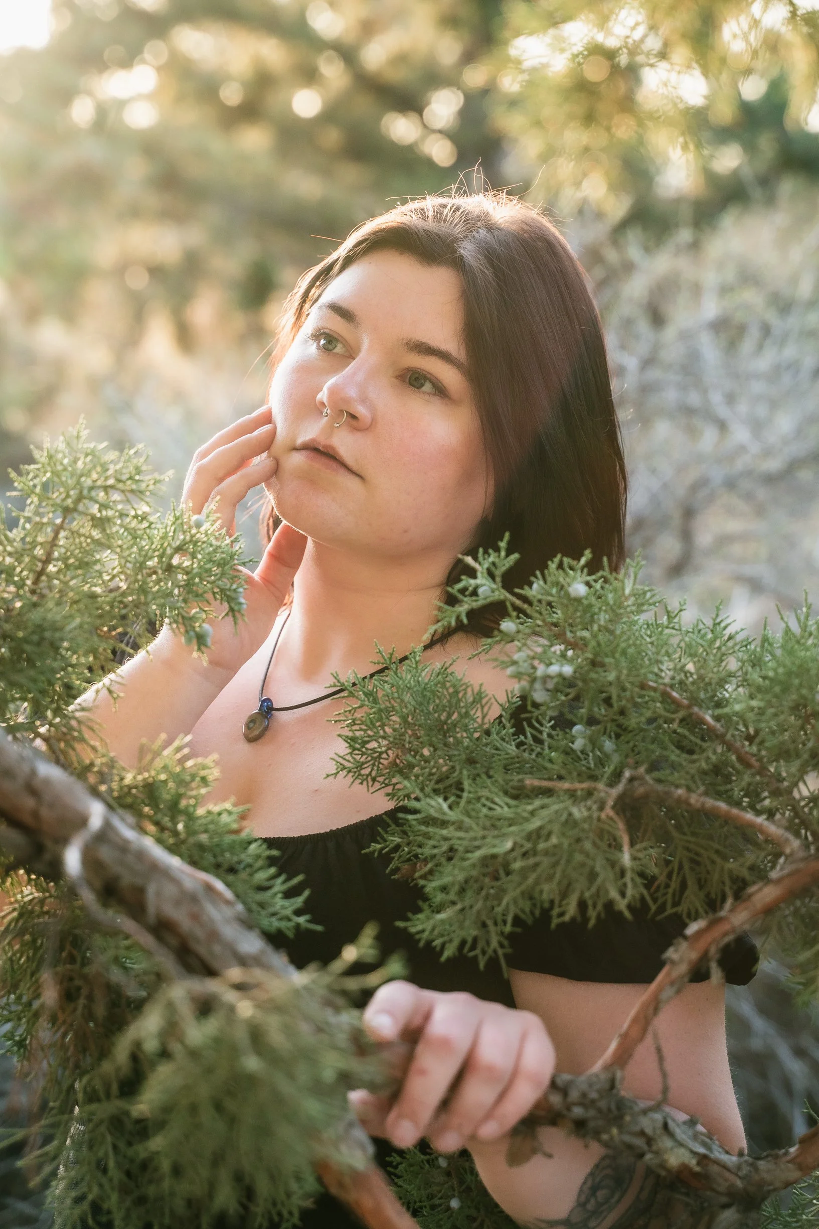 A young woman with short dark hair and a septum piercing standing among trees and greenery, with sunlight filtering through the branches.
