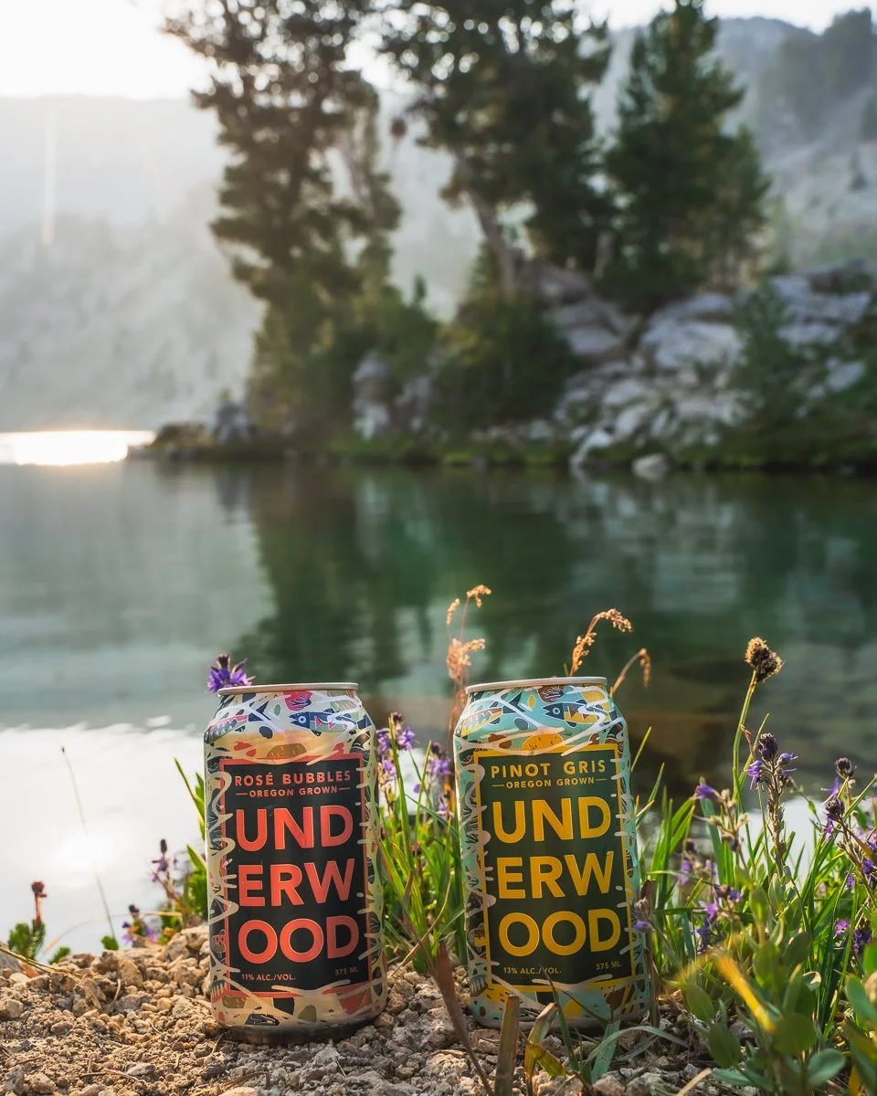 Two cans of Underwood craft beverages, one Rosé Bubbles and one Pinot Gris, placed on the ground among purple flowers near a lake with trees and rocky cliffs in the background, during daylight.