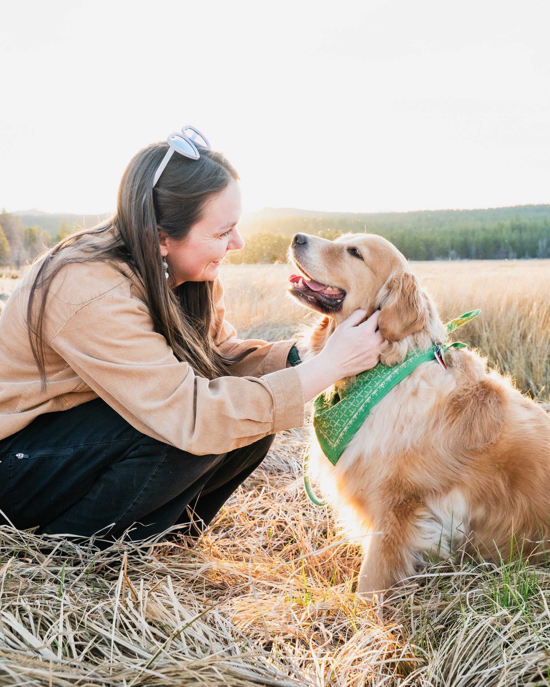 A woman kneeling in a field, smiling and petting a golden retriever dog wearing a green bandana, during sunset.