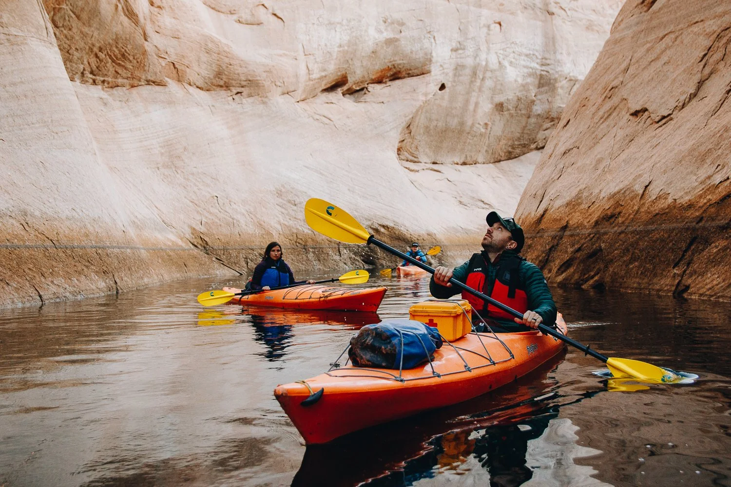 Three people kayaking through a canyon with high, smooth rock walls, wearing life jackets and caps, using paddles to navigate calm waters.