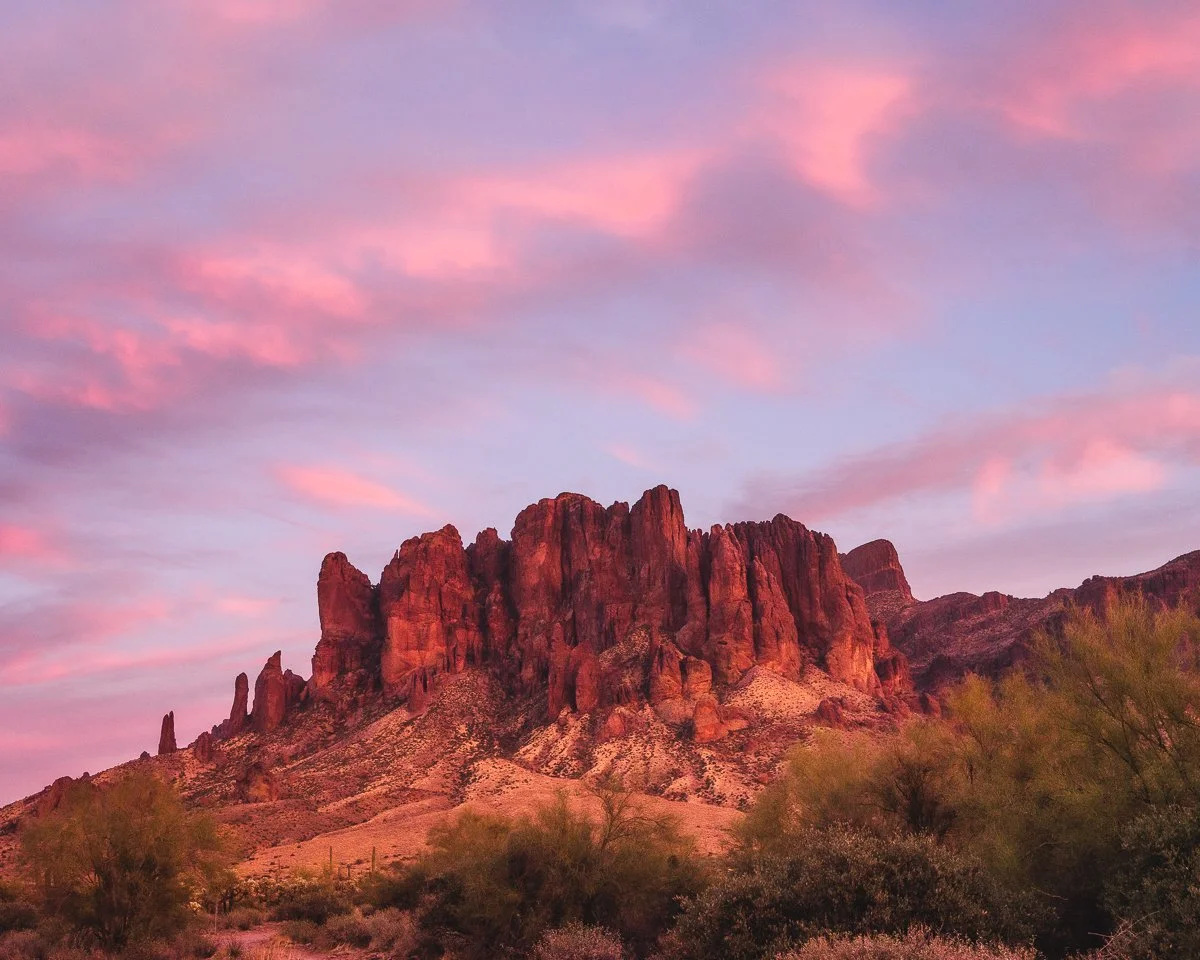 Pink and purple sky over a rugged red rock mountain in a desert landscape with sparse bushes in the foreground.
