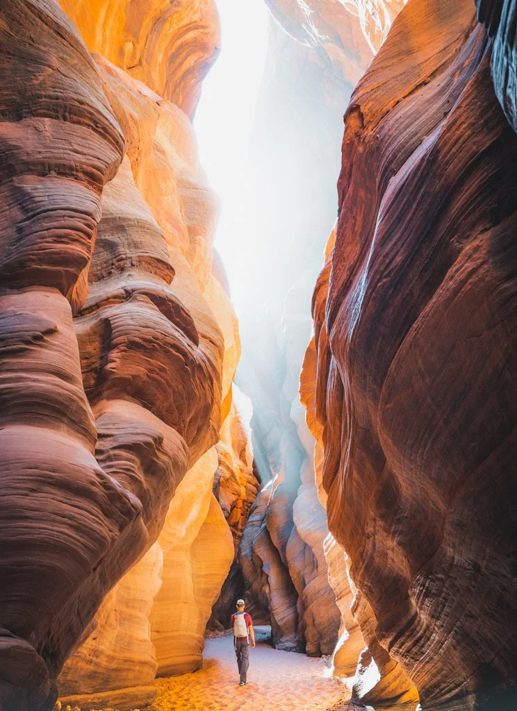 A person walking through a narrow slot canyon with tall, colorful, layered rock walls on either side and sunlight shining down from above.