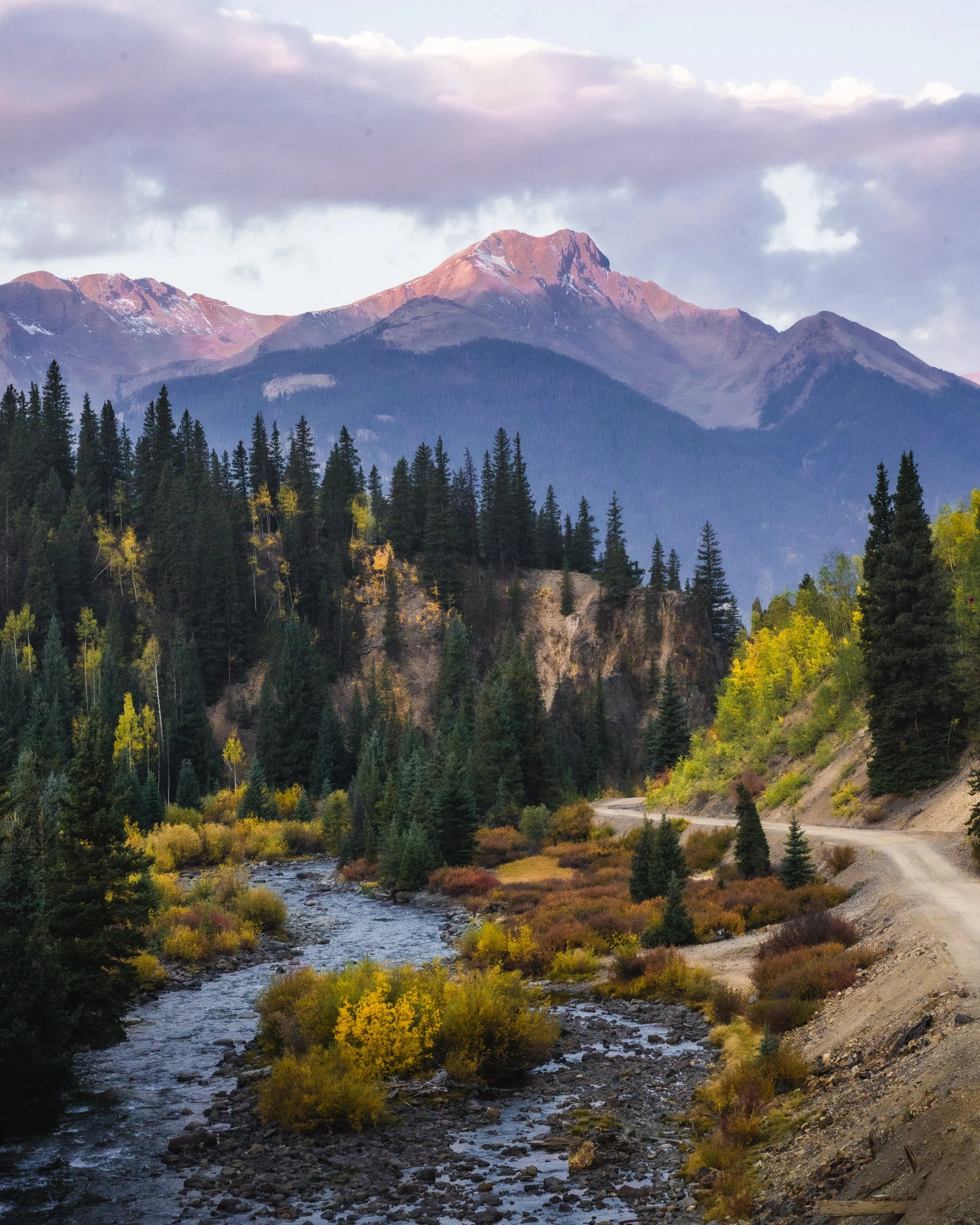 Scenic mountain landscape with a river running through a forested valley and snow-capped peaks in the background.
