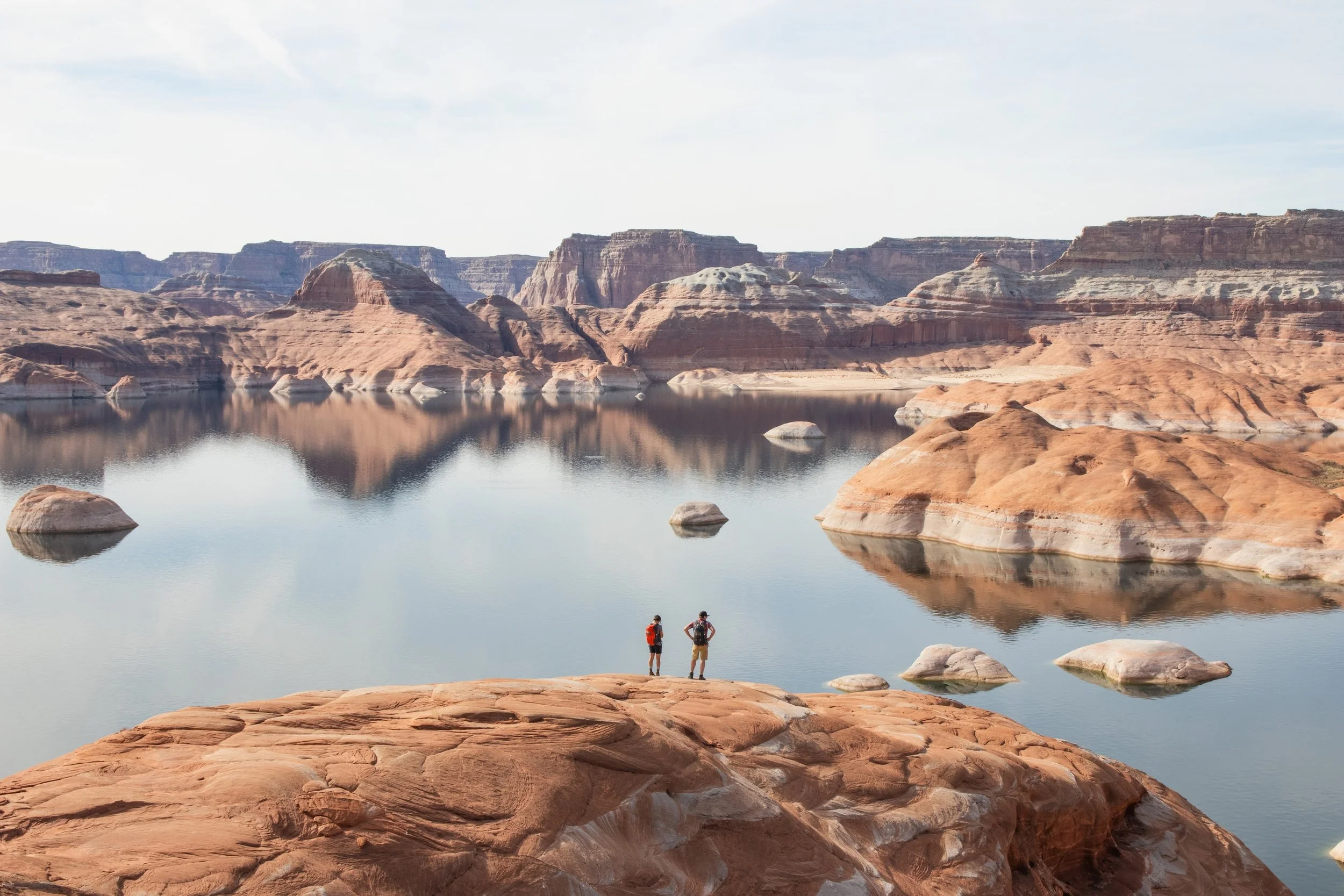 Two hikers in red and black backpacks standing on a rocky ledge overlooking a calm lake with reflections of orange and beige canyon cliffs under a cloudy sky.