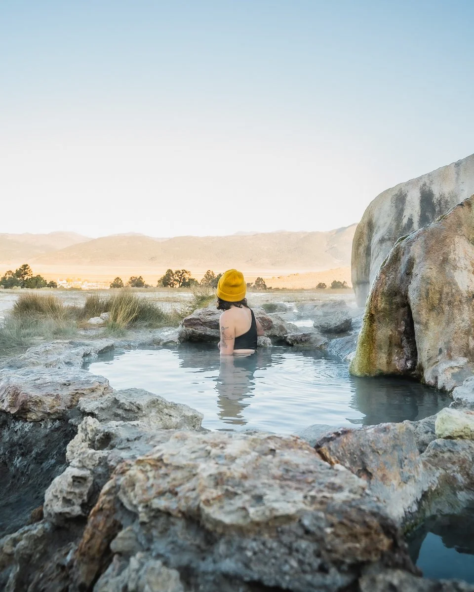 Person with a yellow beanie and tattoos sitting in a hot spring surrounded by rocks with a mountain landscape in the background