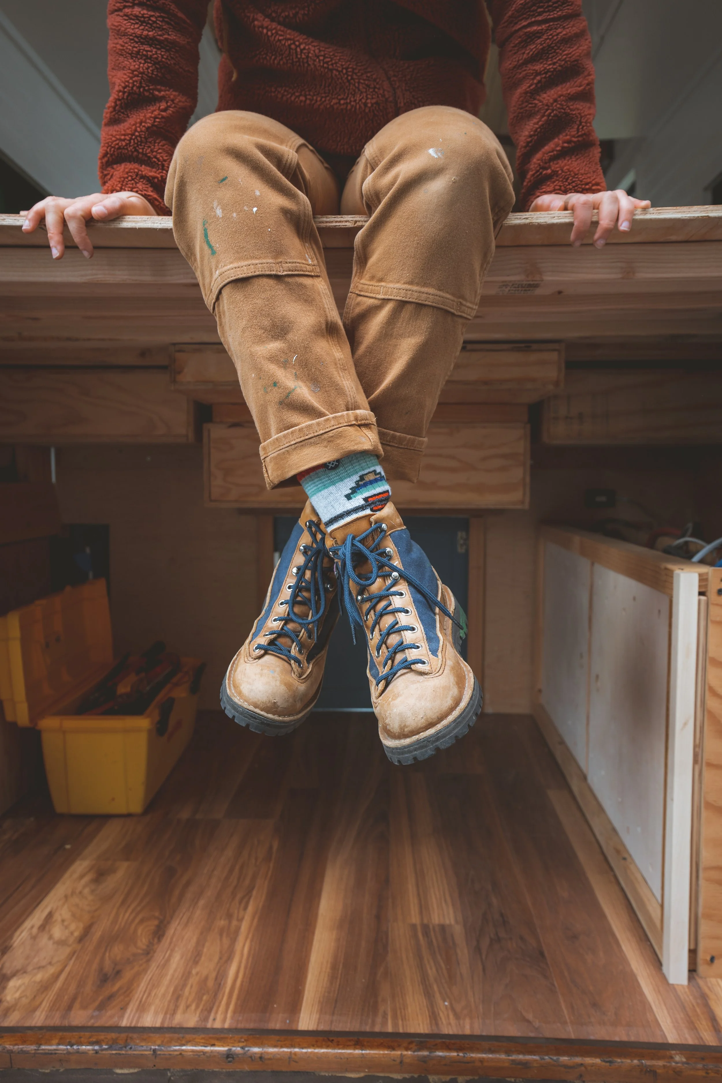 Person wearing tan work boots and colorful socks hanging upside down under a wooden table in a workshop.