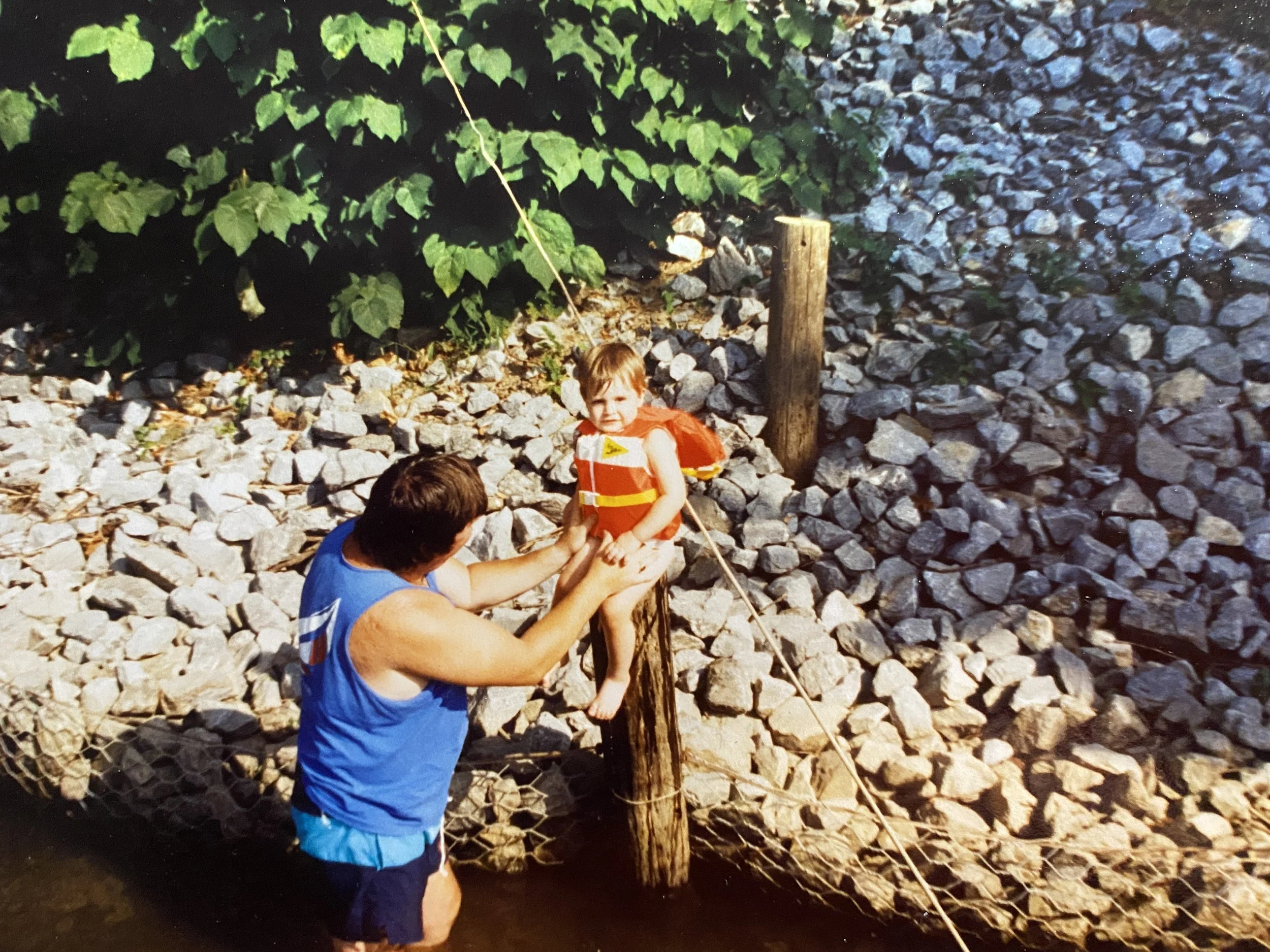 A vintage photo of Craig's grandfather, Jack, holding him up in a lifejacket on a pole by the boat dock along the Potomac River.