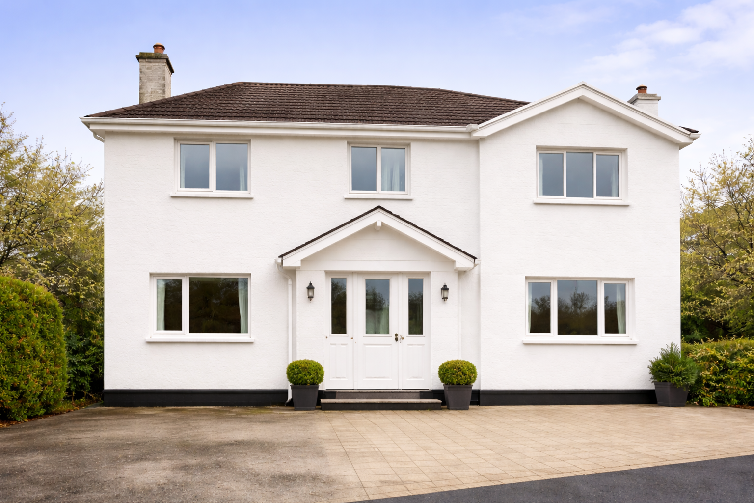 White two-story house with six windows, a double front door, and potted plants near the entrance, with greenery on each side and a paved driveway in front.