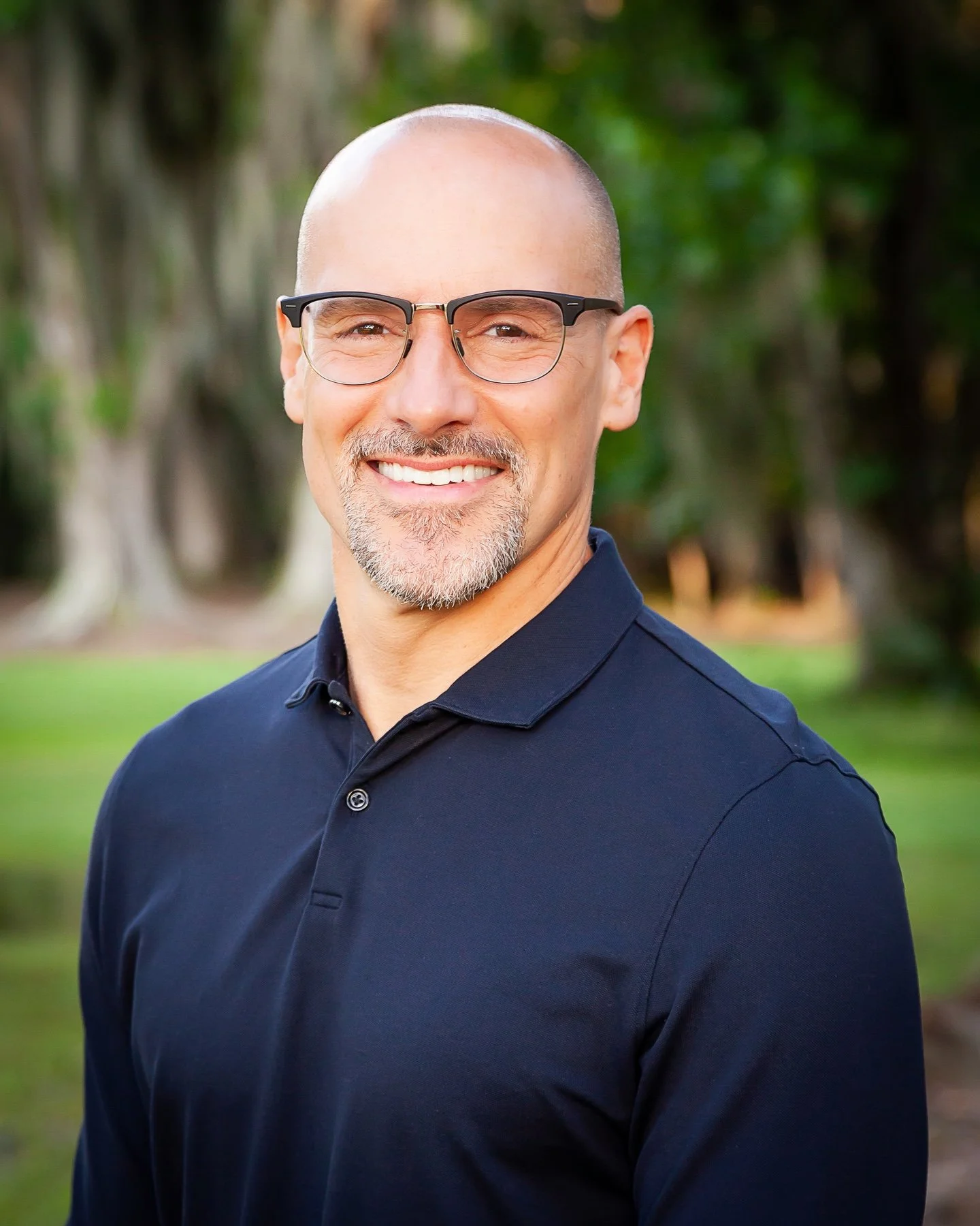 A smiling man with glasses, a goatee, and a shaved head wearing a navy blue polo shirt outdoors with blurred green trees and grass in the background.