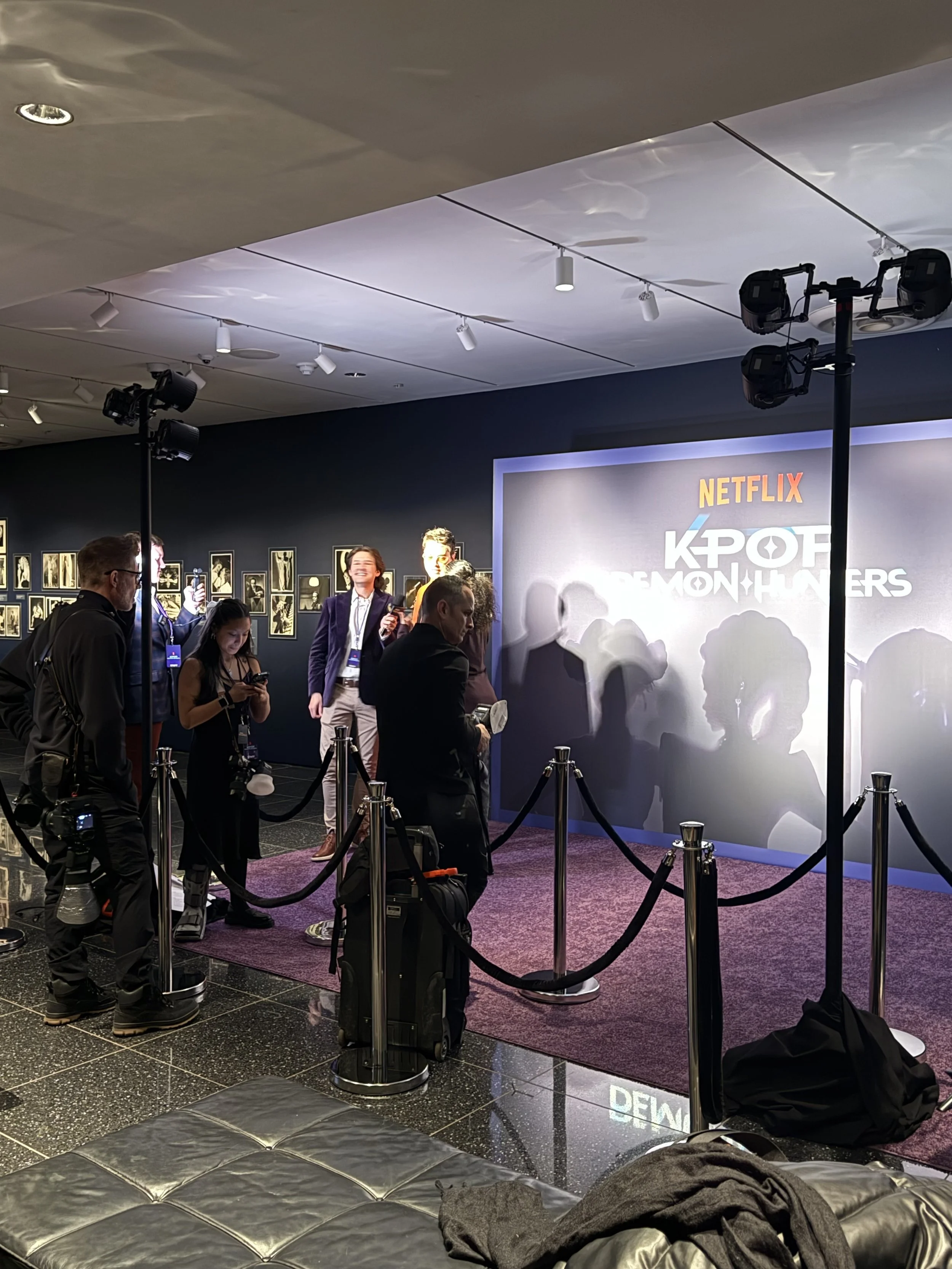 People gathered around a red carpet at a promotional event for 'KOF: Demon Hunters,' sponsored by Netflix. The backdrop features the show's logo, and there are photographers and attendees taking photos, with some waiting behind stanchions.