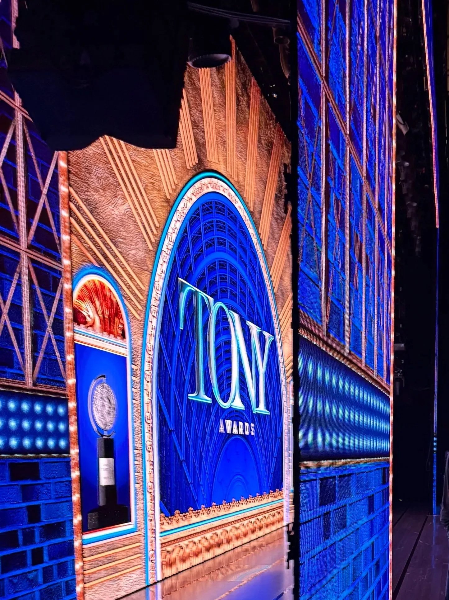 The image shows a brightly lit stage with a large sign reading 'TONY AWARDS' in illuminated letters. The backdrop features an ornate design with gold accents and blue lighting, suggesting an award ceremony setting.