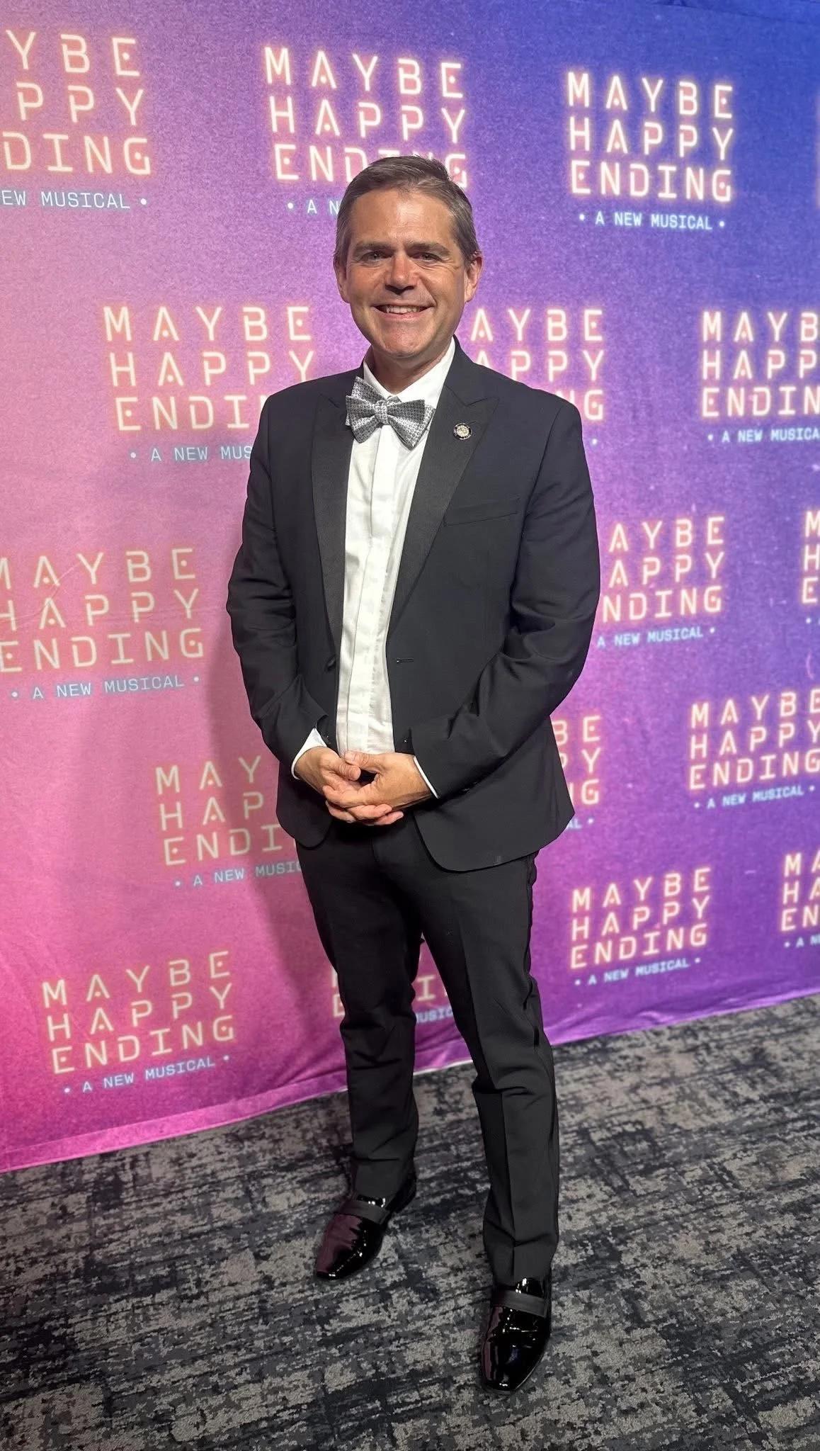 Man in tuxedo with bow tie standing in front of a backdrop with the words 'Maybe Happy Ending' repeated on it.