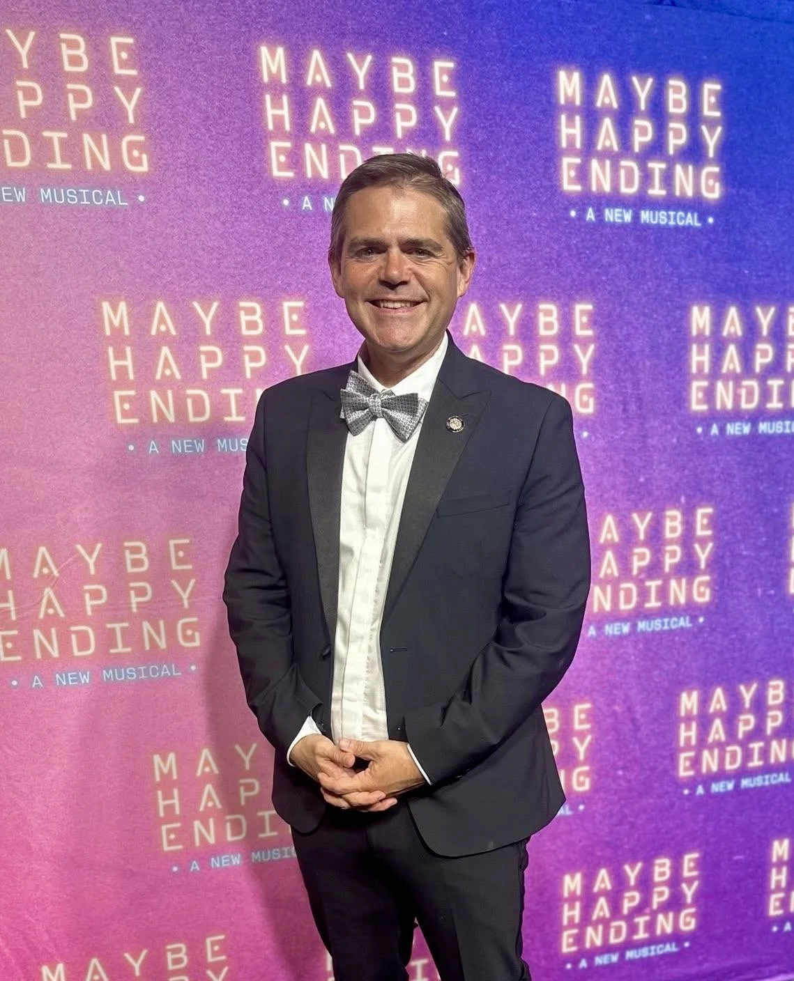 Man in tuxedo with bow tie standing in front of backdrop promoting musical 'Maybe Happy Ending.'