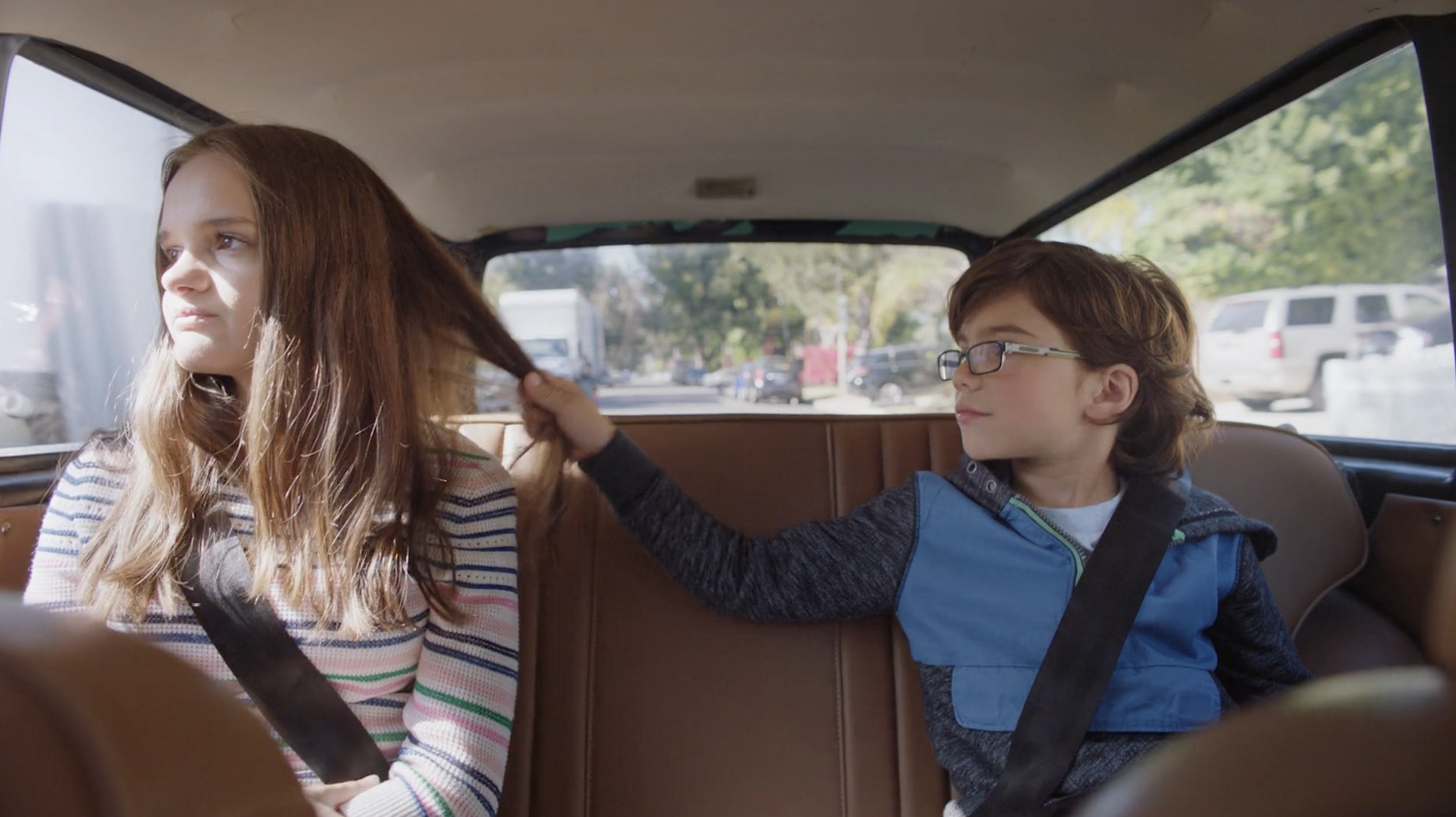 A young girl and boy sitting in the backseat of a car, the girl looking out the window with a serious expression and the boy playfully pulling her hair.