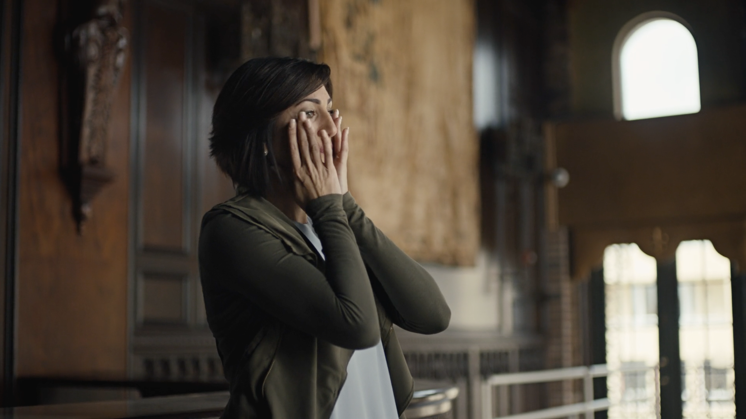 A woman with short dark hair covering her face with her hands, standing inside a rustic wooden building near large windows.