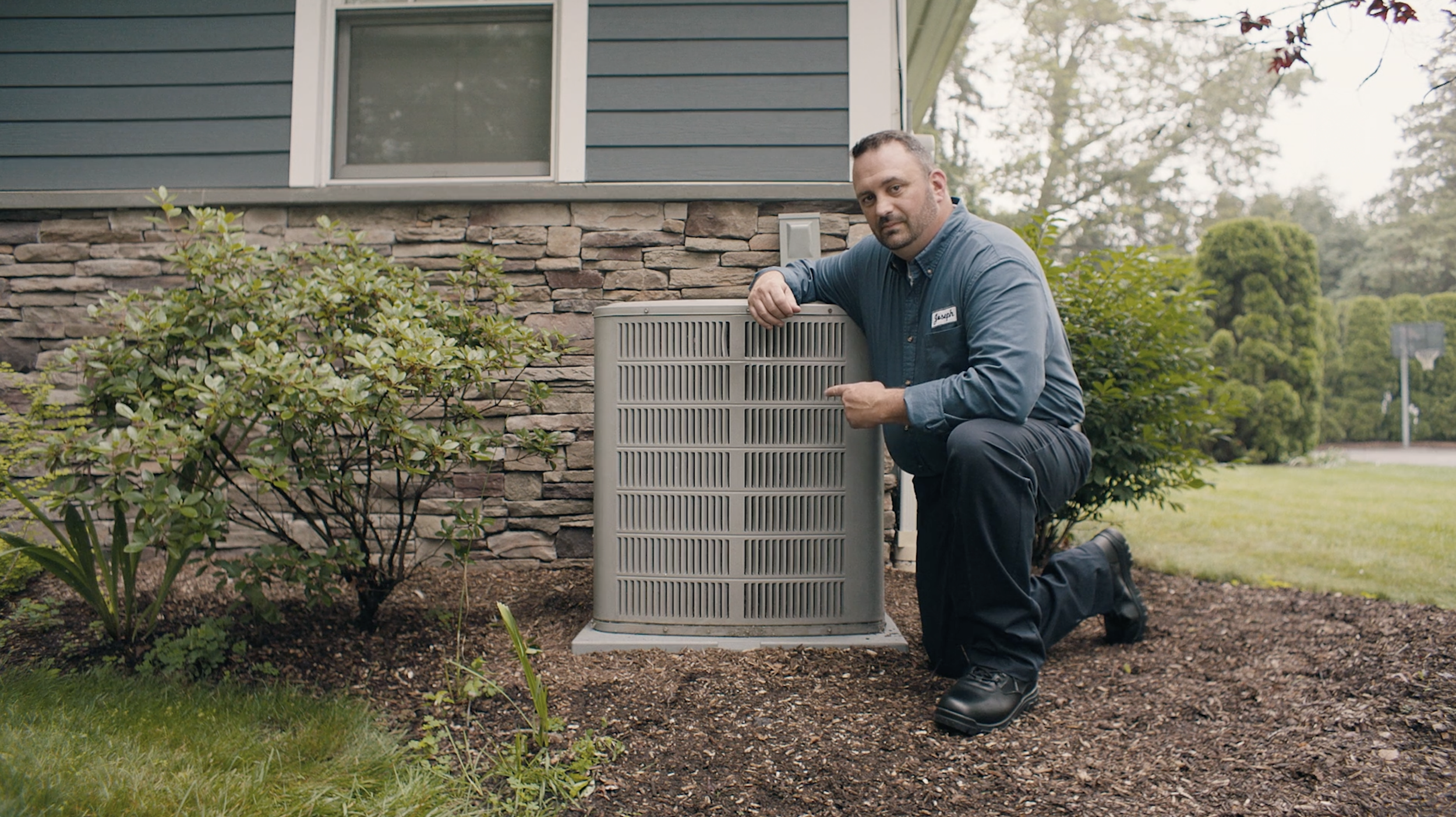 A man crouching next to an outdoor air conditioning unit on a lawn in front of a house with stone and siding exterior.