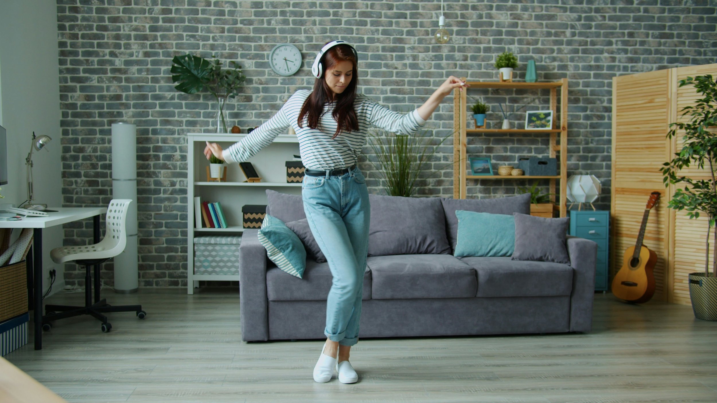 Young woman dancing in living room, wearing headphones, striped shirt, and light jeans, with a sofa, bookshelf, and guitar in the background.