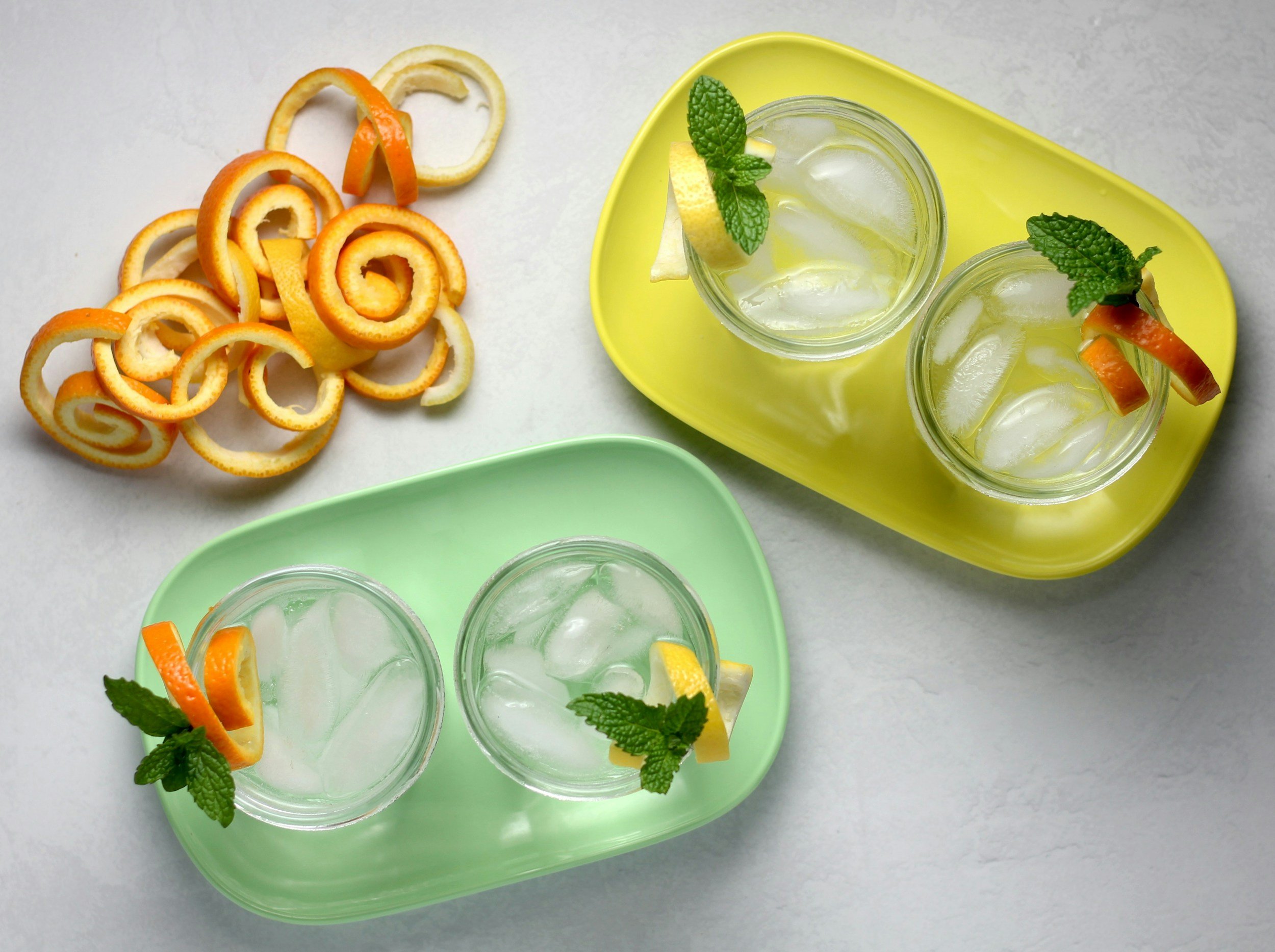 Two glasses of lemonade with ice, lemon slices, and mint leaves on colorful trays, alongside lemon and orange peels.