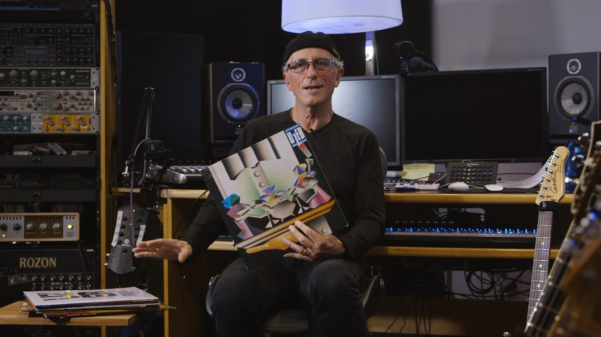 Un homme parlant dans un studio d'enregistrement ou de production musicale, assis devant une console de mixage avec plusieurs ordinateurs, haut-parleurs et équipements audio.