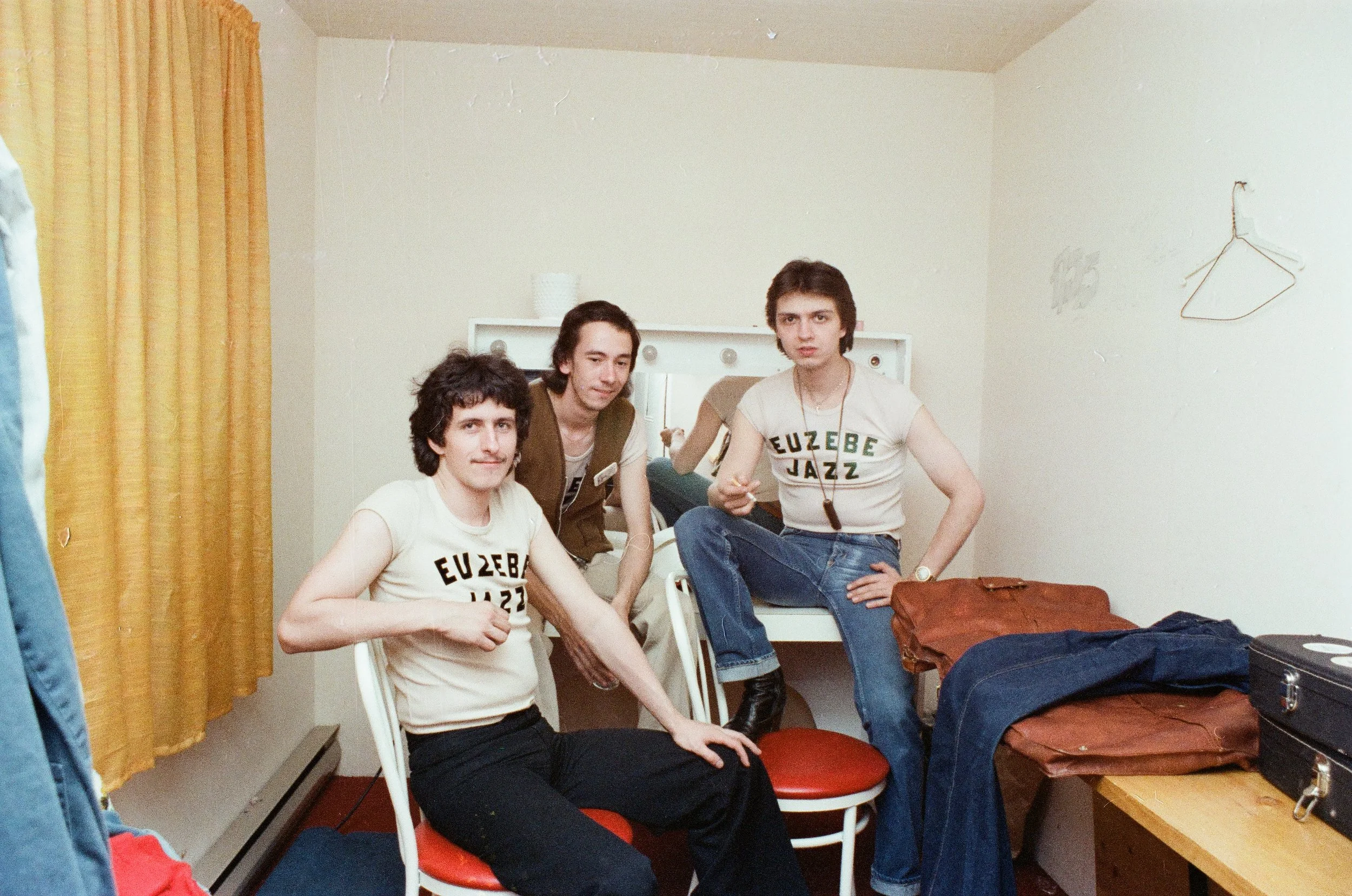 Trois jeunes hommes posent dans une chambre avec des vêtements sur une table.