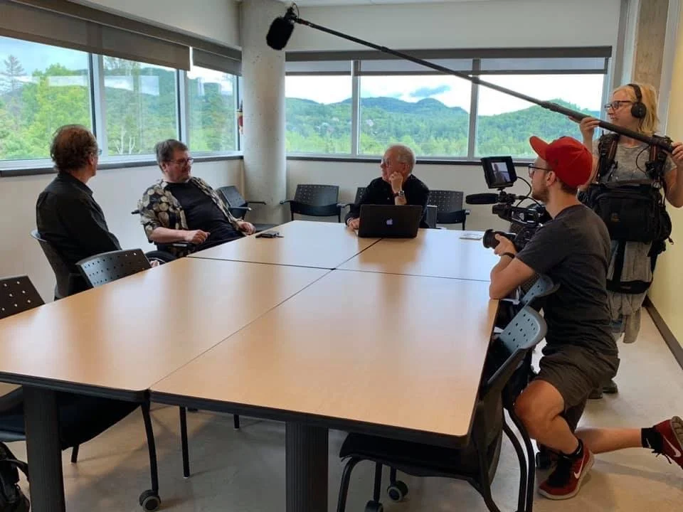 Une équipe de tournage filme un entretien lors d'une interview dans une salle de réunion. Un homme avec une casquette rouge est assis, un autre avec un ordinateur portable est également présent, ainsi que deux autres personnes à gauche. La personne à droite tient un microphone directionnel.