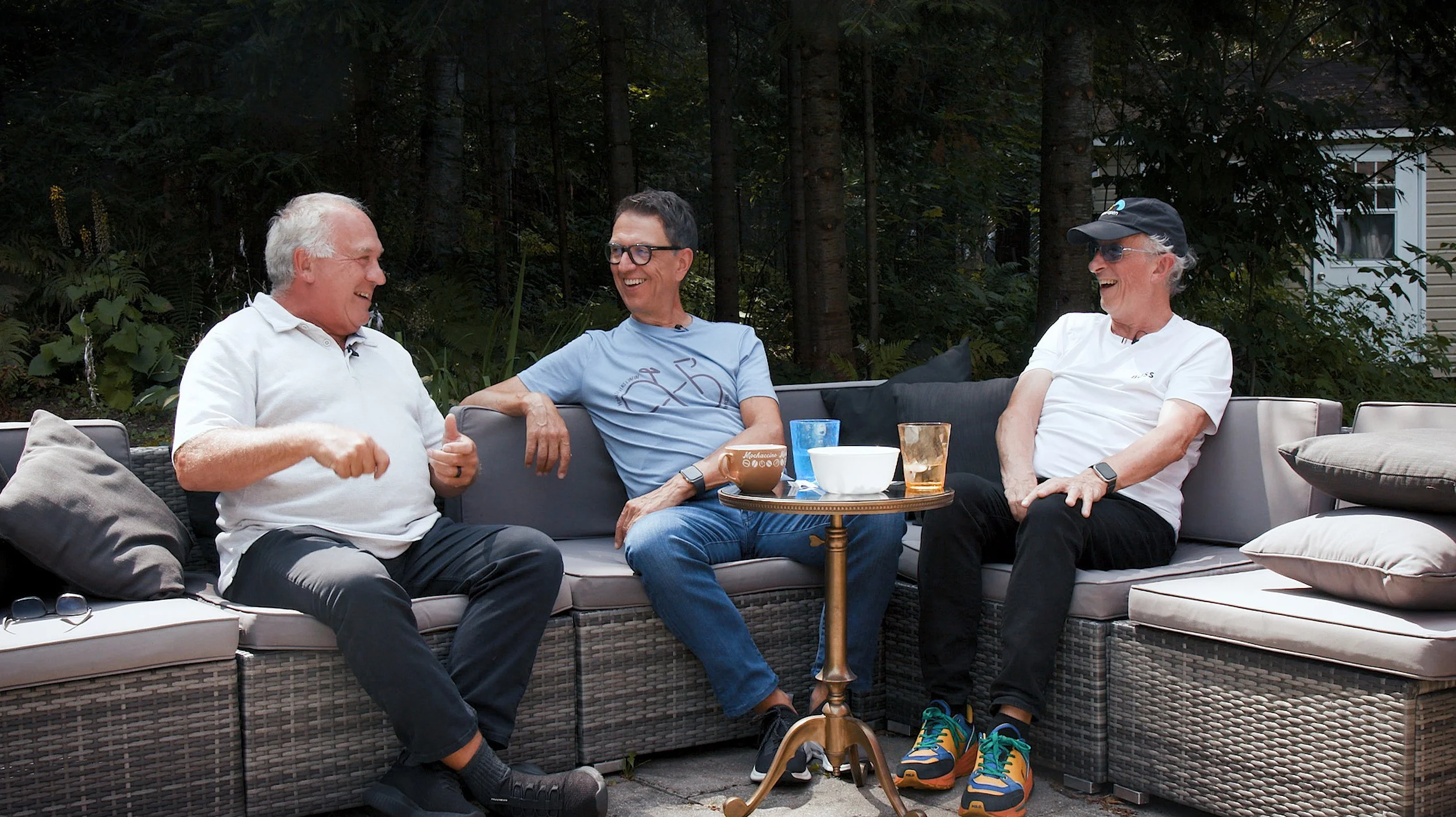 Trois hommes âgés assis sur un canapé en plein air, en train de rire et discuter, avec une table basse devant eux contenant des verres et des bols, entourés de végétation dense.