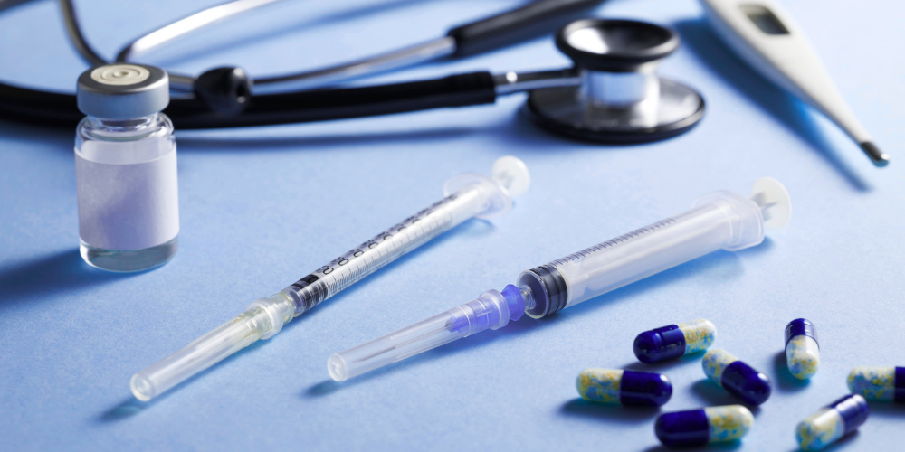 Medical equipment including syringes, capsules, a stethoscope, a thermometer, and a small vial on a blue surface.