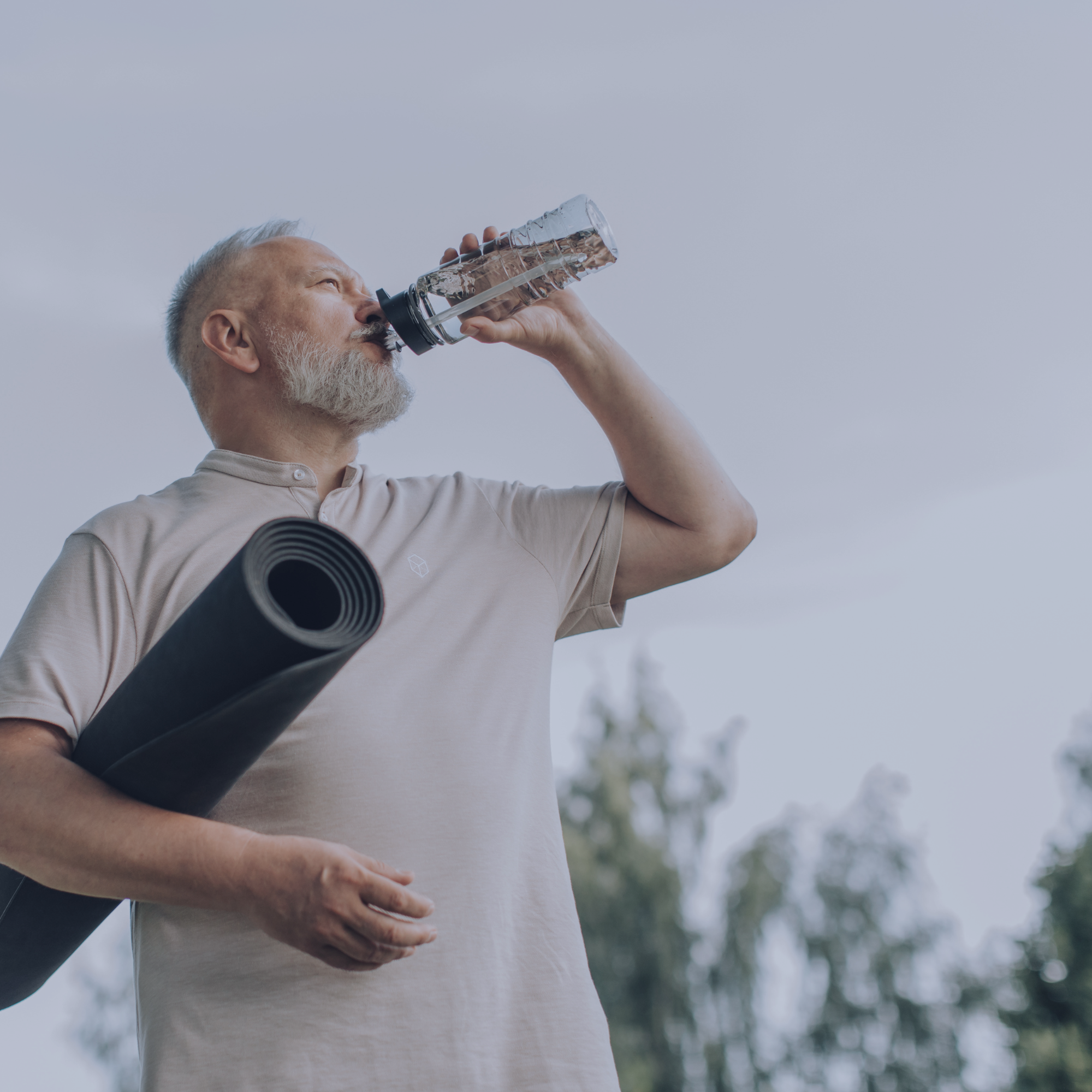 An older man with gray hair and a beard drinks water from a clear plastic water bottle outdoors. He wears a beige polo shirt and is holding a rolled-up yoga mat under his arm with trees and sky in the background.