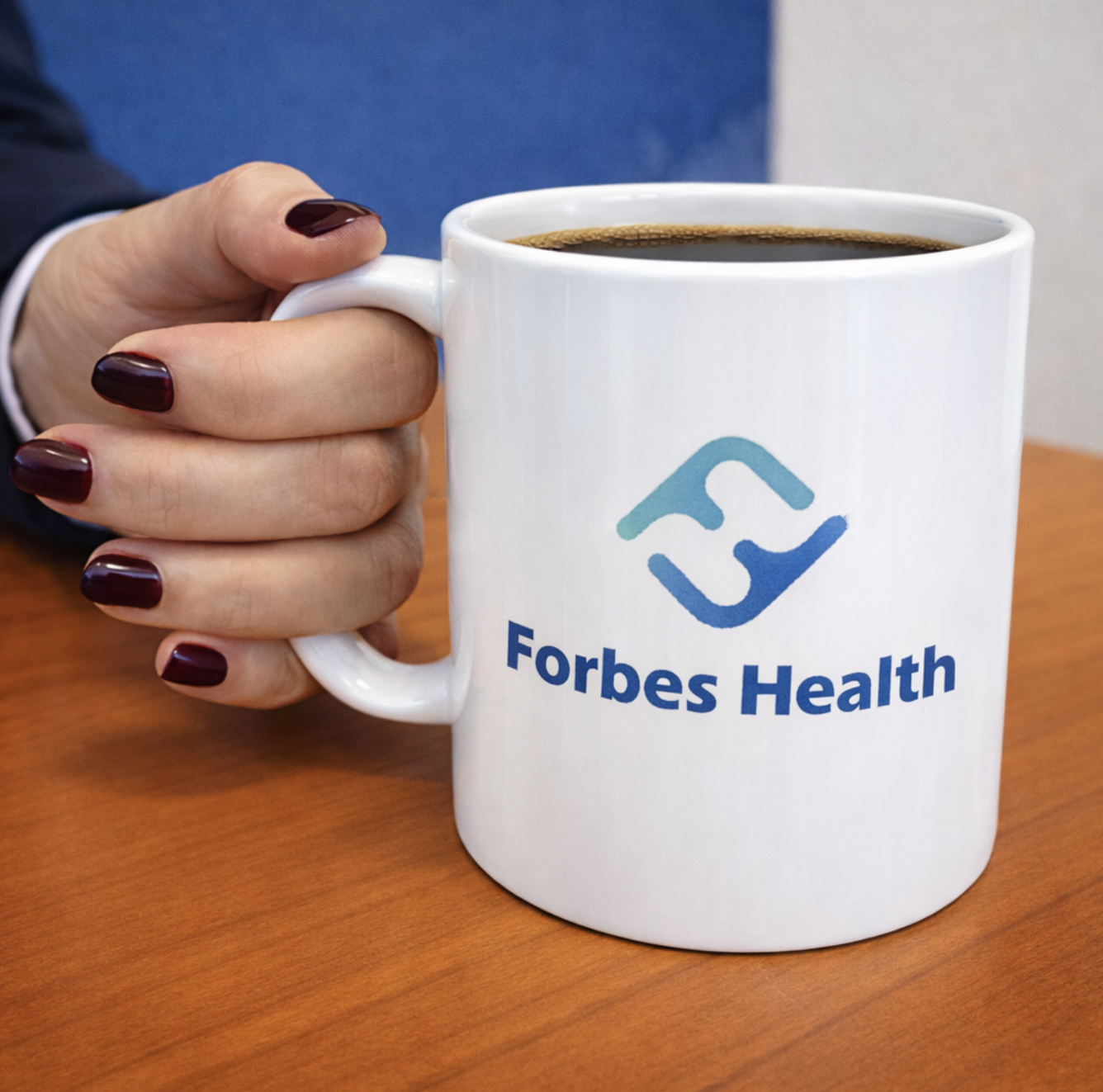 A person's hand holding a white coffee mug with the Forbes Health logo, filled with black coffee, on a wooden table.