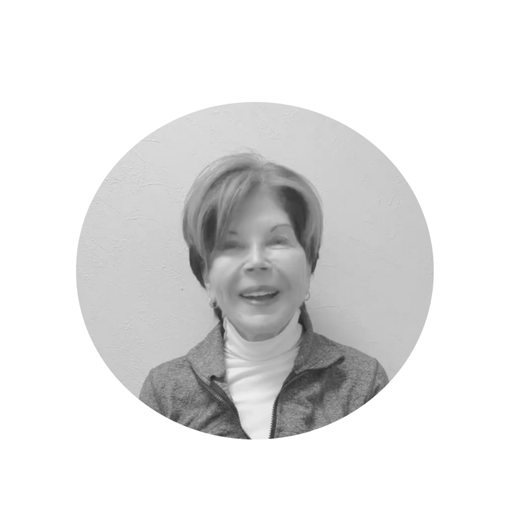 Black and white photo of a smiling woman with short hair, wearing a zip-up jacket and a white turtleneck, against a plain wall.