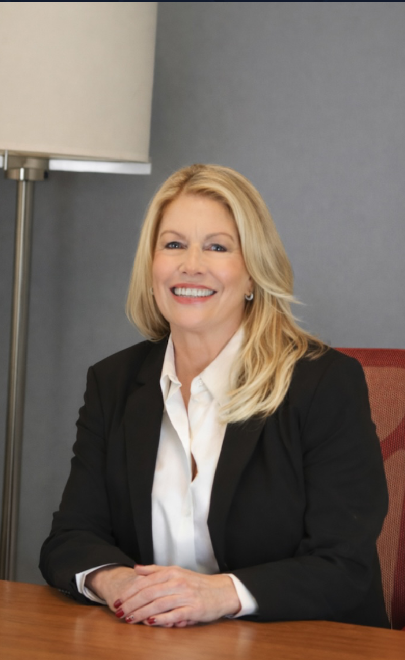 A woman with blonde hair smiling, sitting at a desk with a lamp in the office, wearing a black blazer and white shirt.