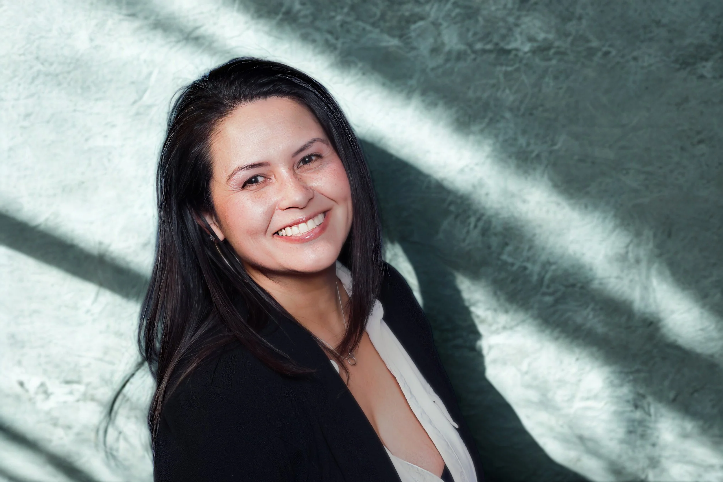 A woman with long dark hair smiling, wearing a black blazer and white top, with sunlight and shadows on a textured green wall background.