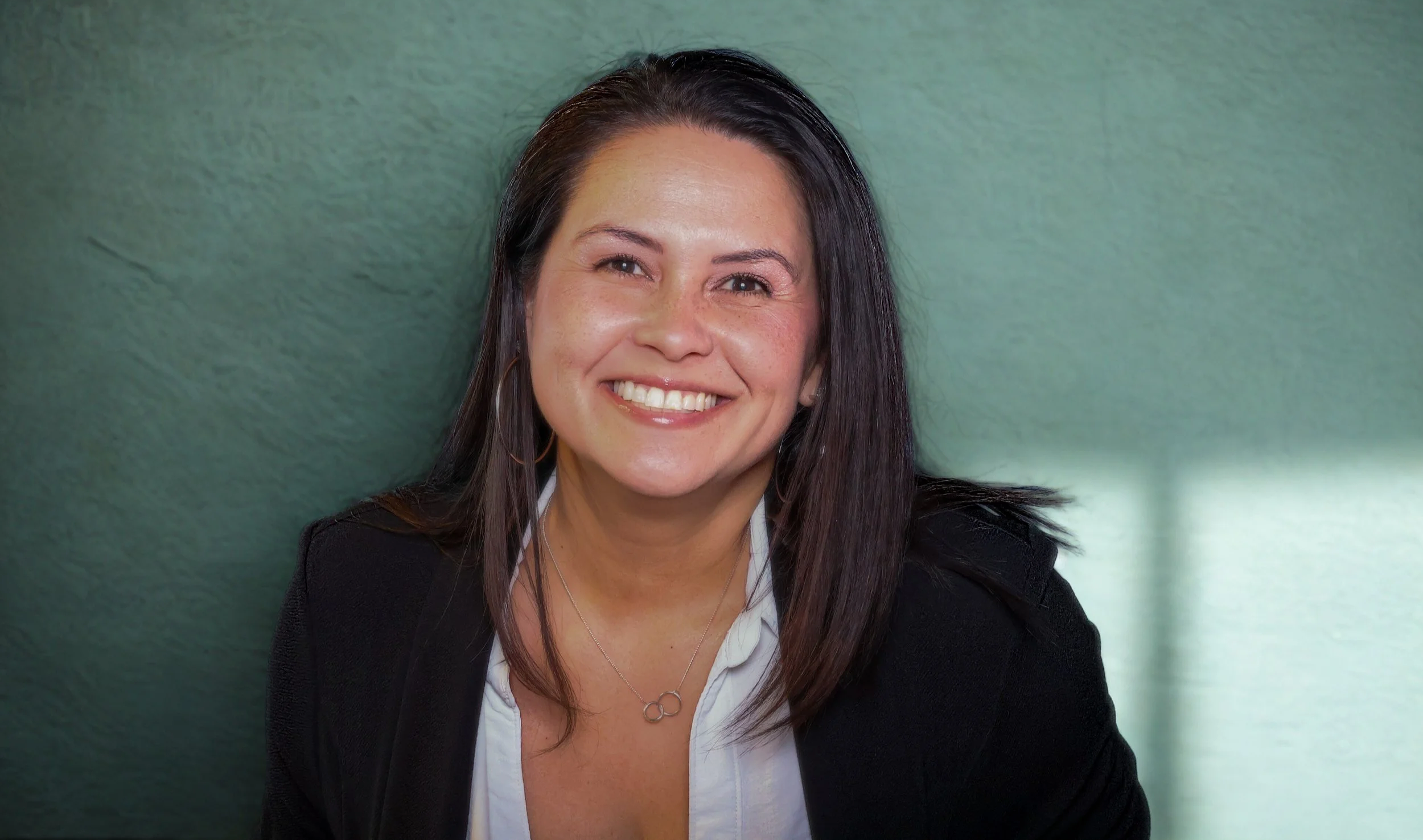 A smiling woman with dark brown hair, wearing a white shirt, black blazer, and a necklace with three interconnected circles, standing against a greenish wall.