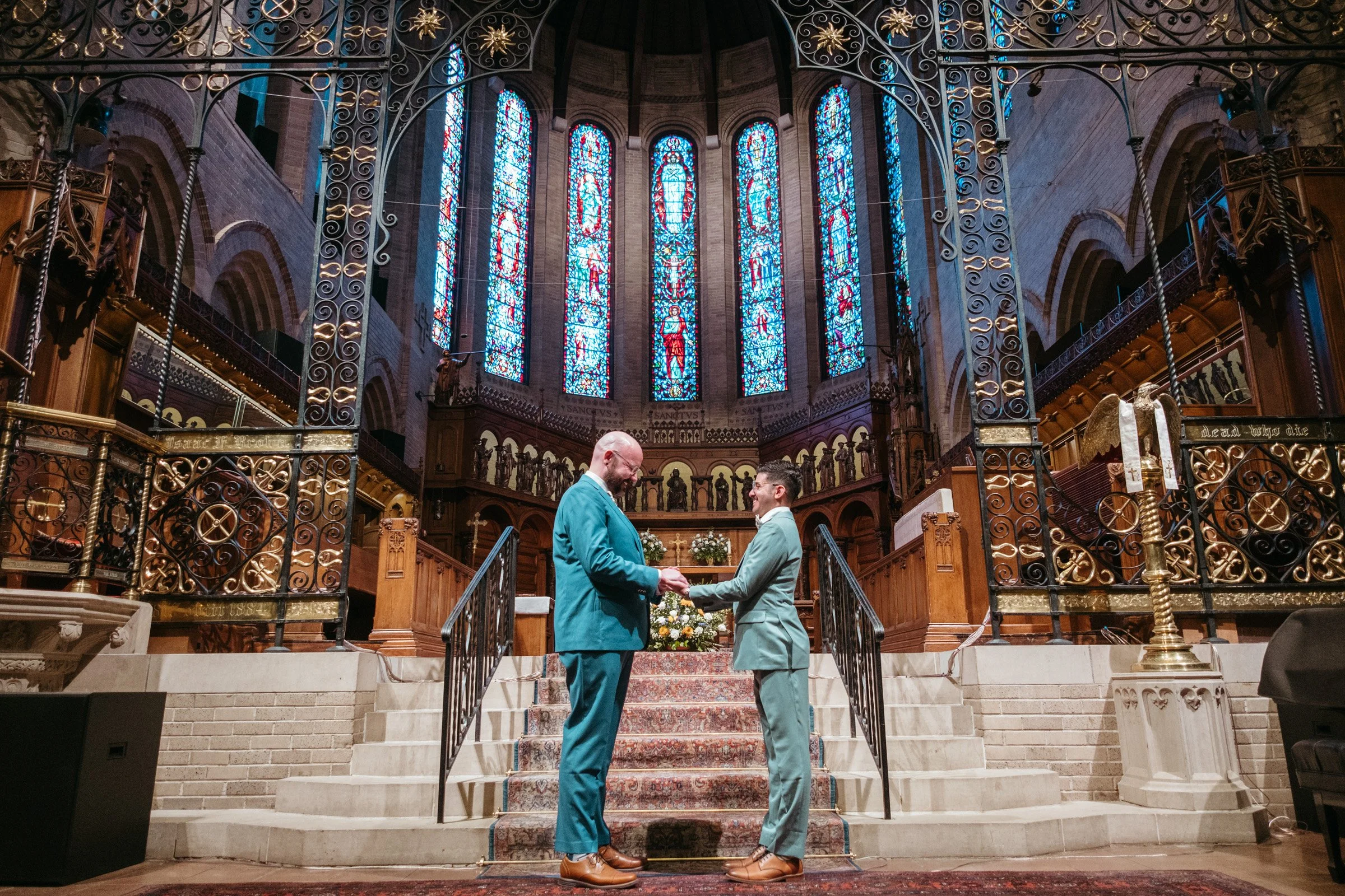 Two men in suits are exchanging rings inside a church with stained glass windows and ornate wooden architecture.