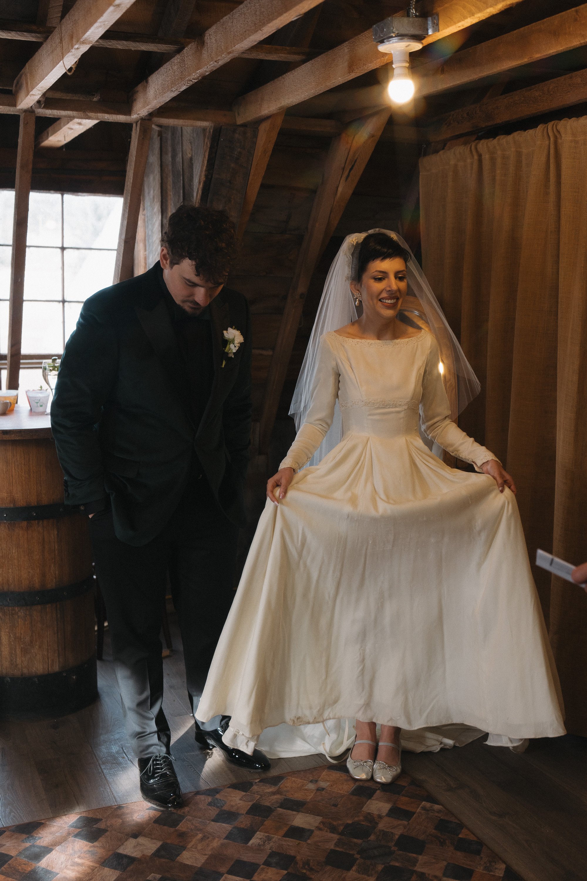 A bride in a white wedding gown and veil, smiling, holding out her dress in a rustic wooden room, standing next to a man in a black suit.